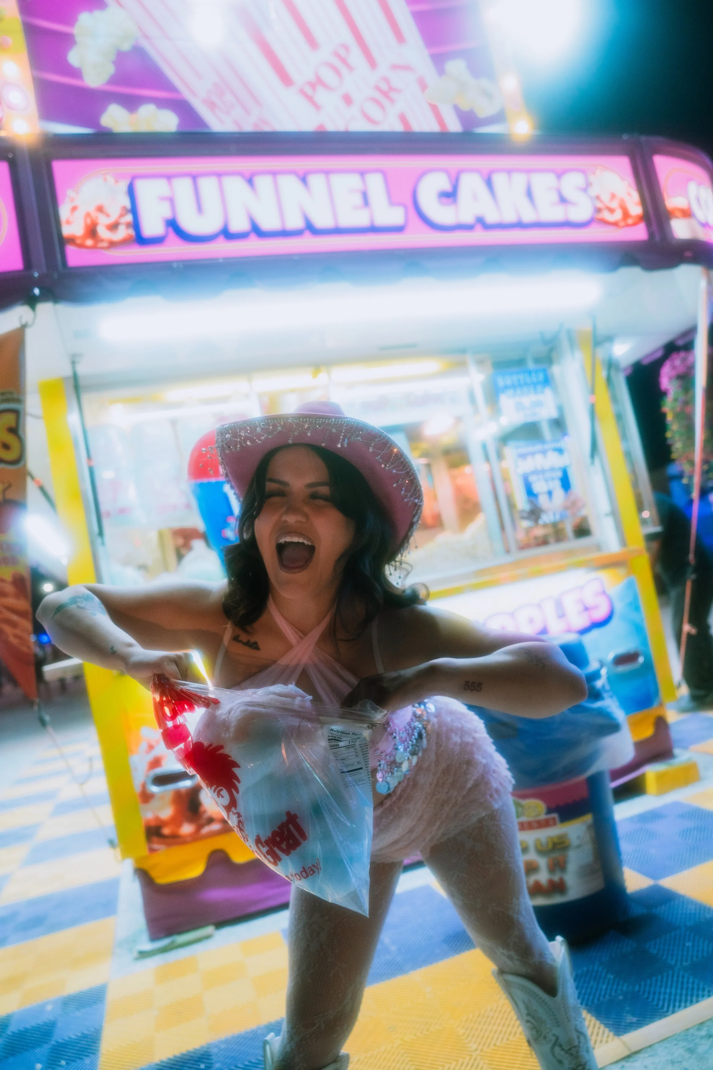A woman wearing a pink hat and a white lace outfit is holding a bag of cotton candy in front of a brightly lit funnel cake stand at a fair or carnival, with the woman appearing to be laughing or shouting in excitement.
