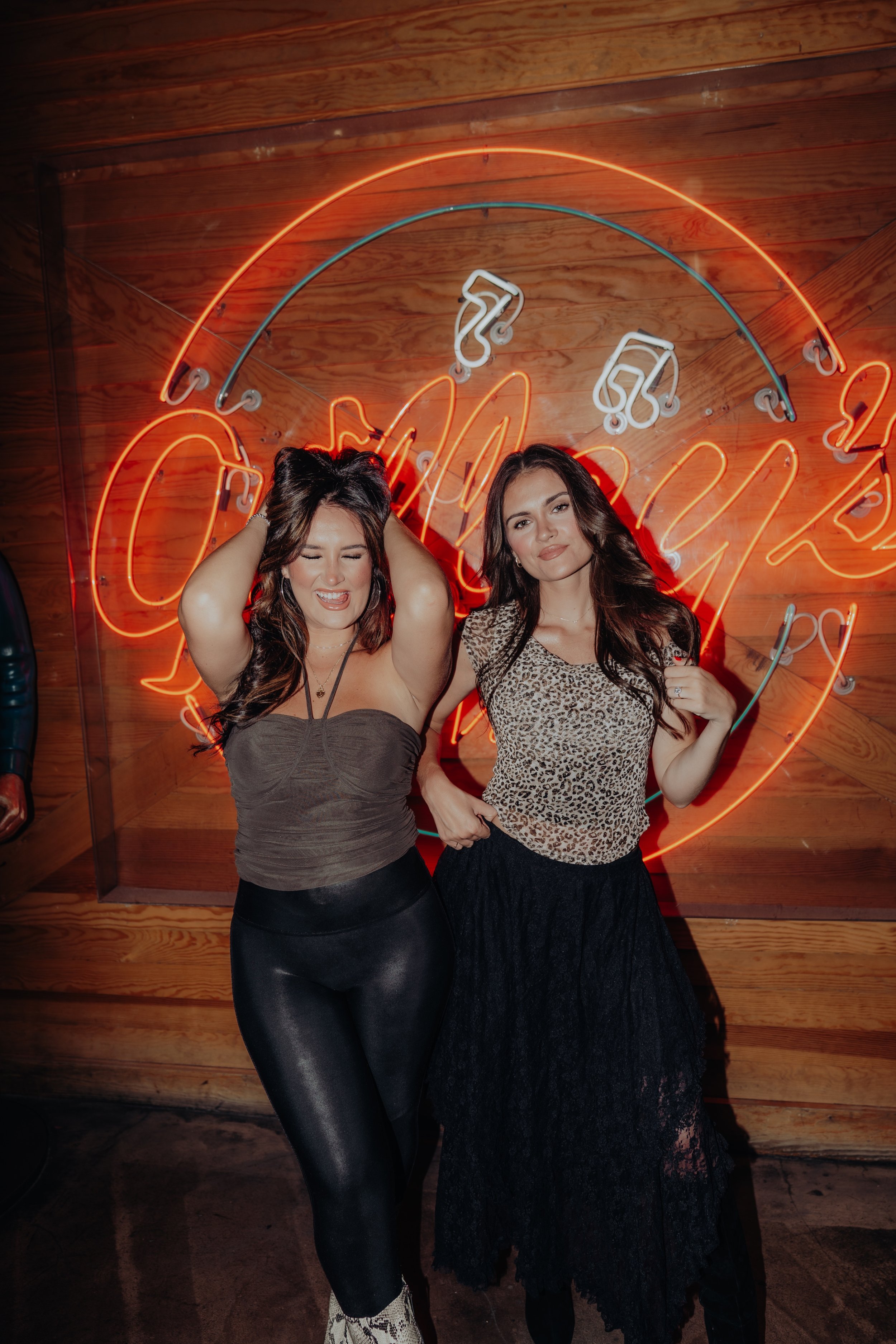 Two women standing in front of a neon sign with a wooden wall background. One woman has her hands behind her head, wearing a strapless top and black pants. The other woman is holding a cigarette, wearing a leopard print top and black lace skirt.