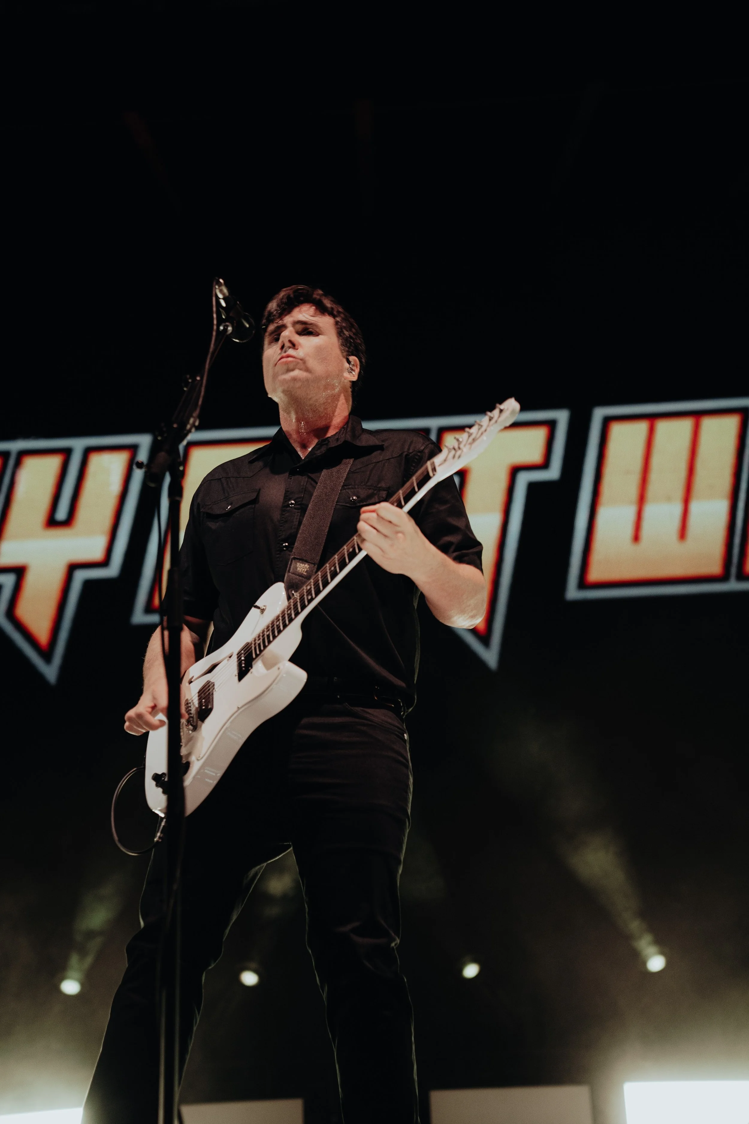 A man with dark hair and a serious expression is playing an electric guitar on stage with a dark background and large illuminated text behind him, illuminated by stage lights.