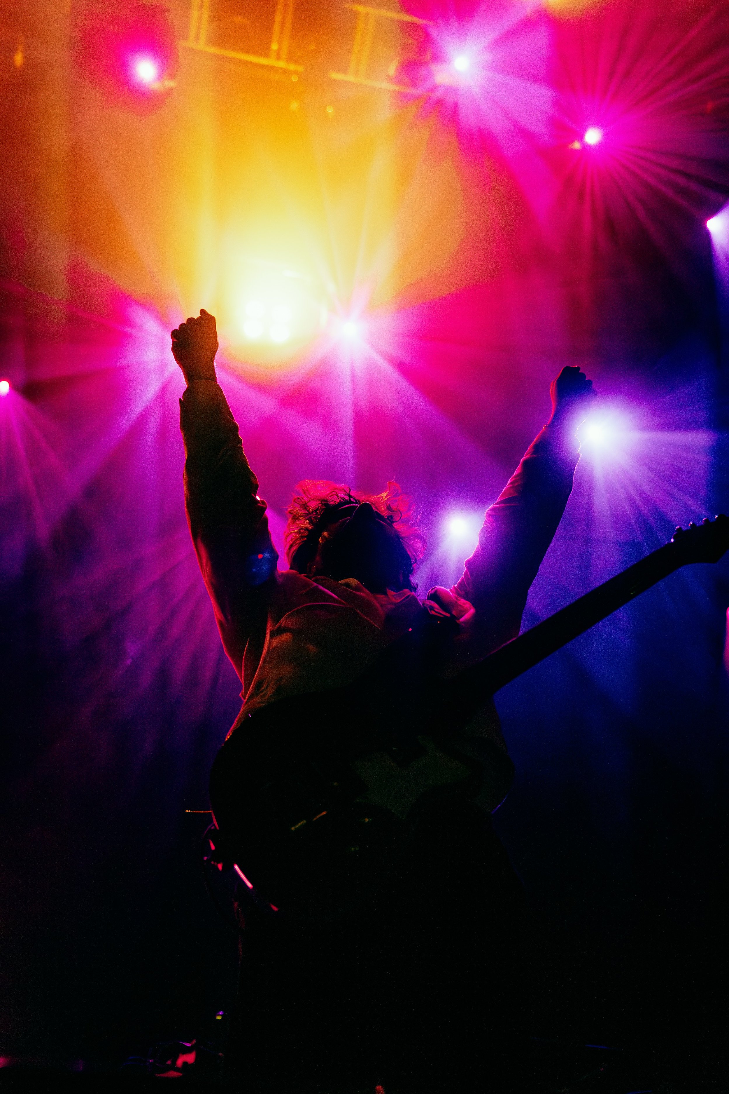 Person with curly hair raising their arms in front of colorful stage lights, including pink, yellow, and purple.
