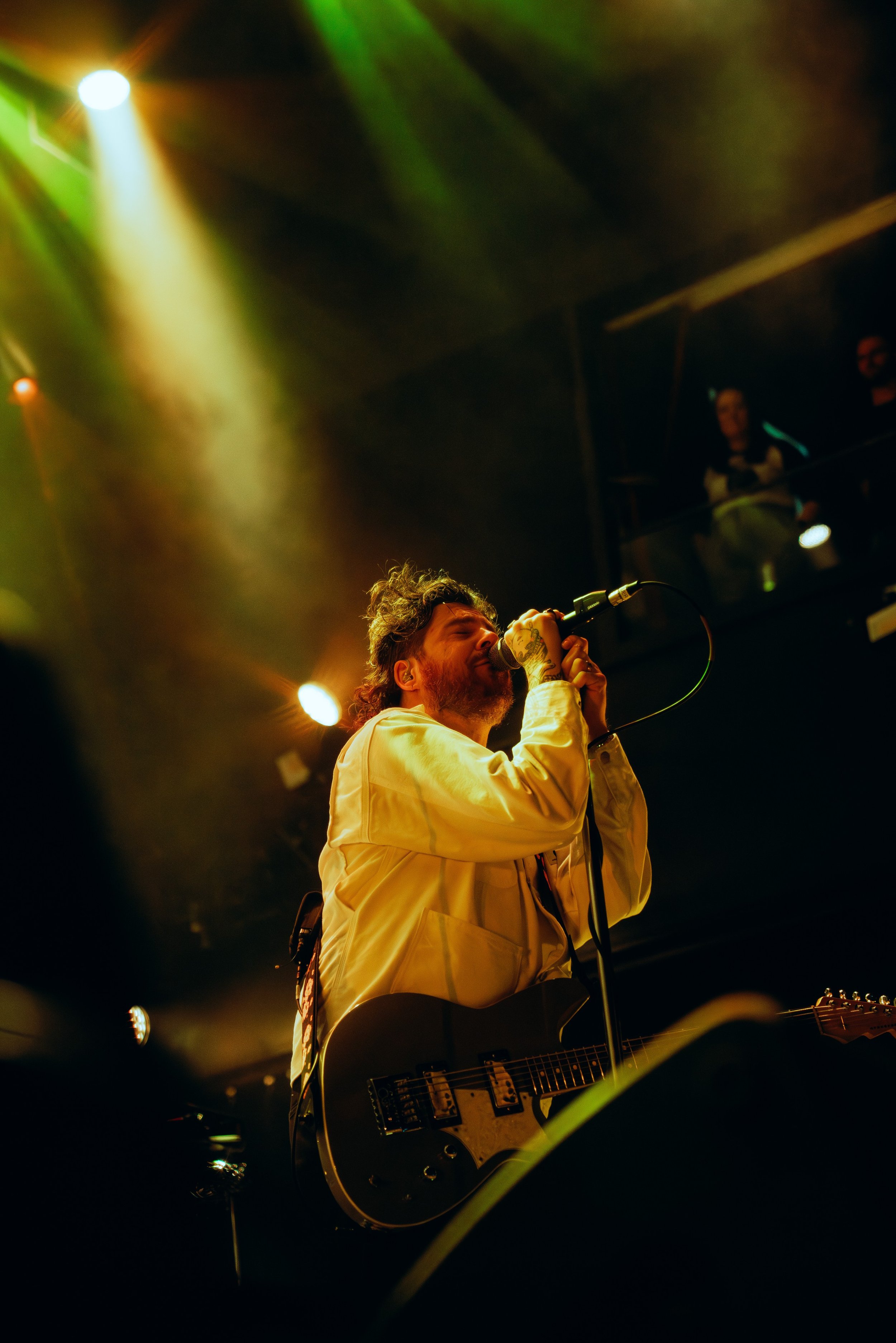 A male singer with dark curly hair and tattoos on his hand, wearing a light-colored jacket, is performing on stage with a microphone and guitar under colorful stage lights, with an audience and a DJ or musician in the background.