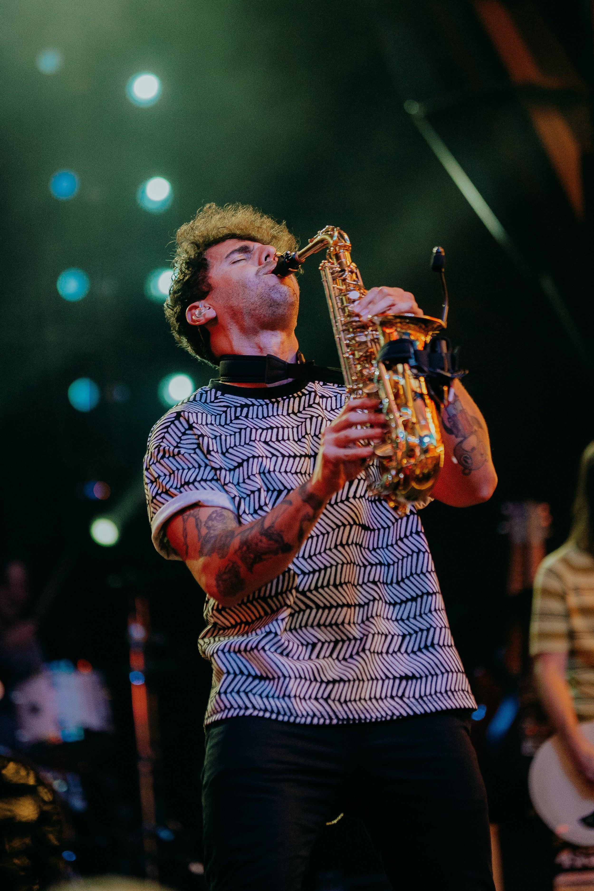A musician playing a gold saxophone on stage, eyes closed, wearing a patterned shirt and tattoos on arm, with colorful stage lights in the background.