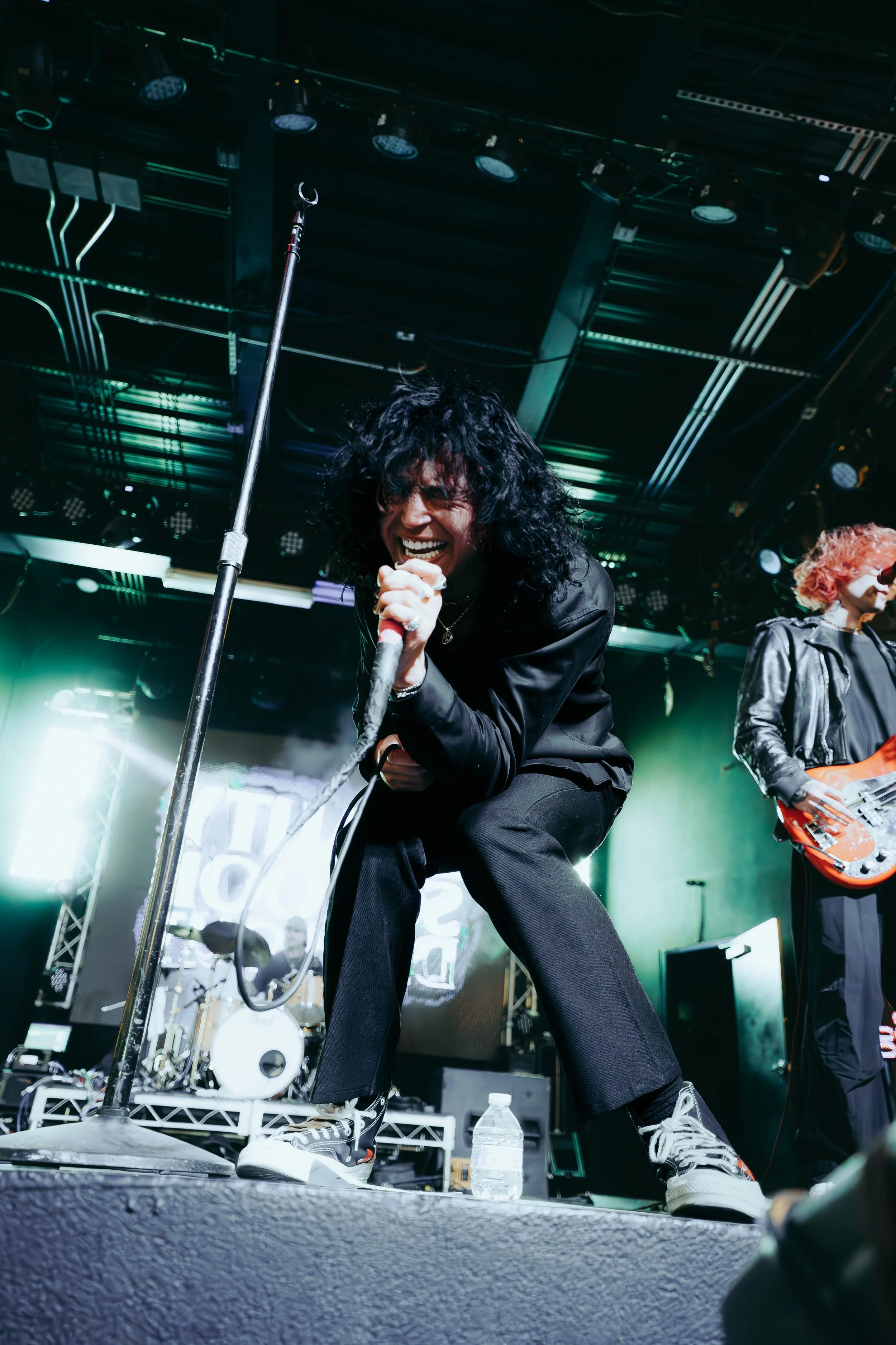 A live music performer with curly black hair singing into a microphone on stage, wearing a black jacket and pants, with a guitarist to their right and stage lights above.