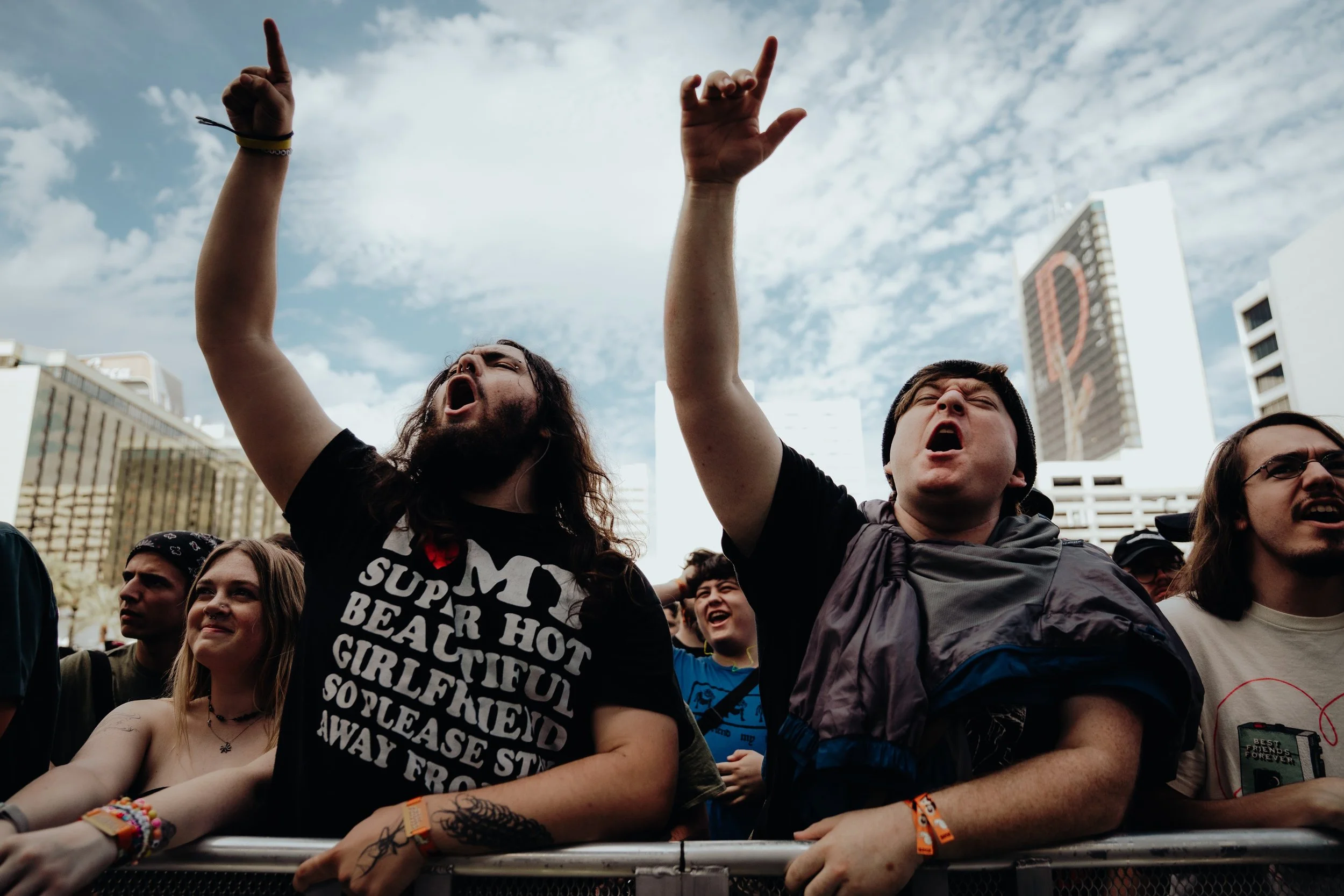 Group of people at an outdoor concert or rally, raising their hands and cheering, with skyscrapers and a partly cloudy sky in the background.