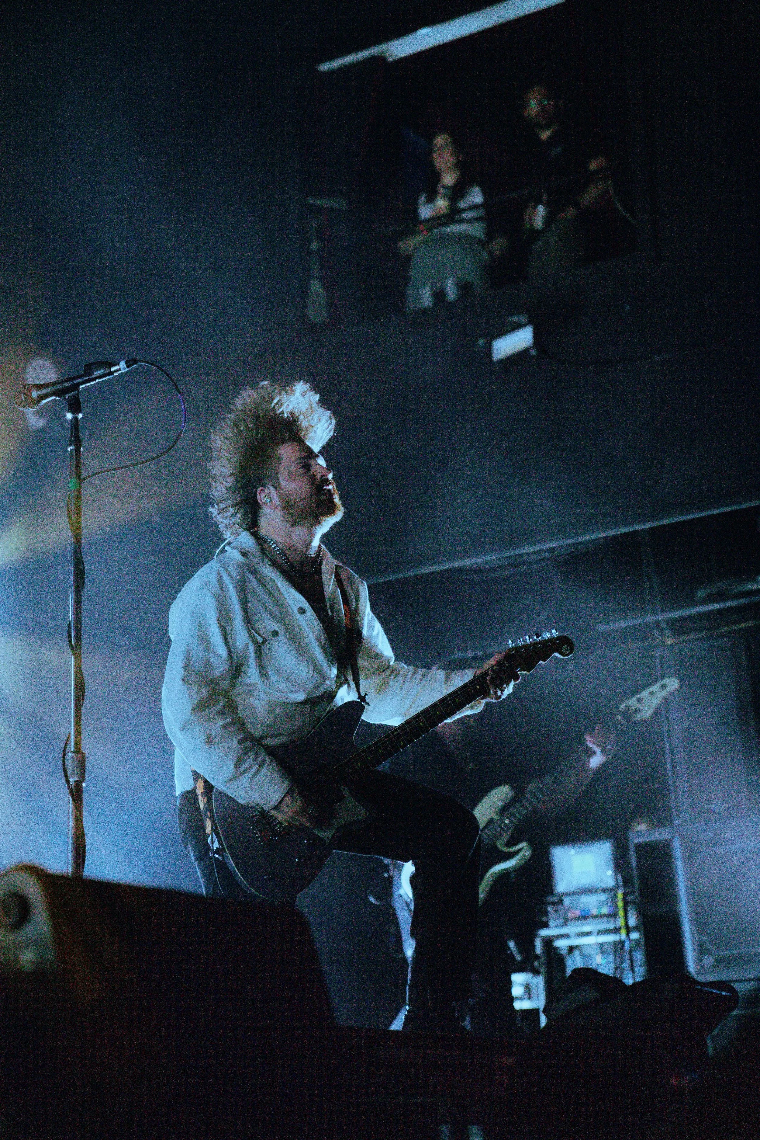 A male musician with a distinctive hairstyle, playing an electric guitar on stage with a microphone stand in front of him and stage equipment visible in the background.