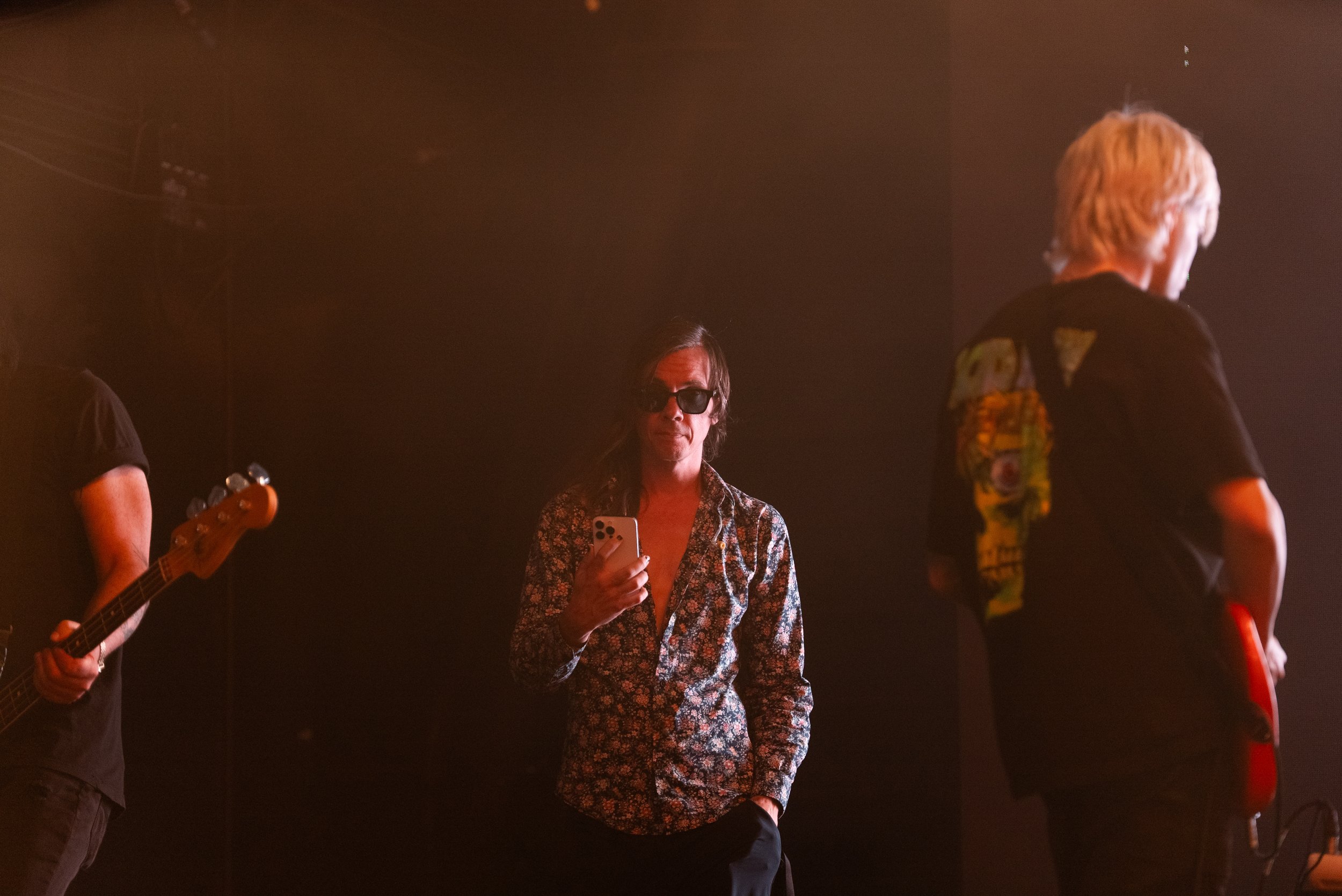 A woman with dark hair and sunglasses holding a smartphone on stage with band members, one playing guitar. The scene is illuminated with warm, reddish stage lighting.