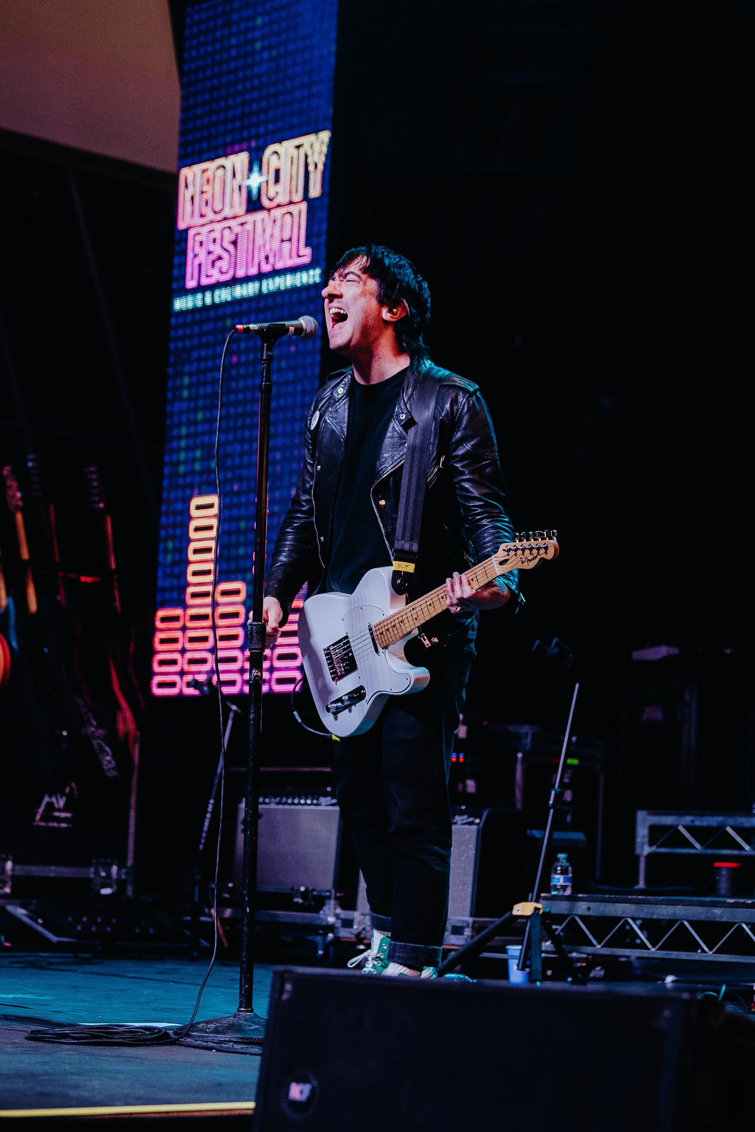 A male musician singing into a microphone while playing an electric guitar on stage at Neon City Festival, with neon-lit signage in the background.