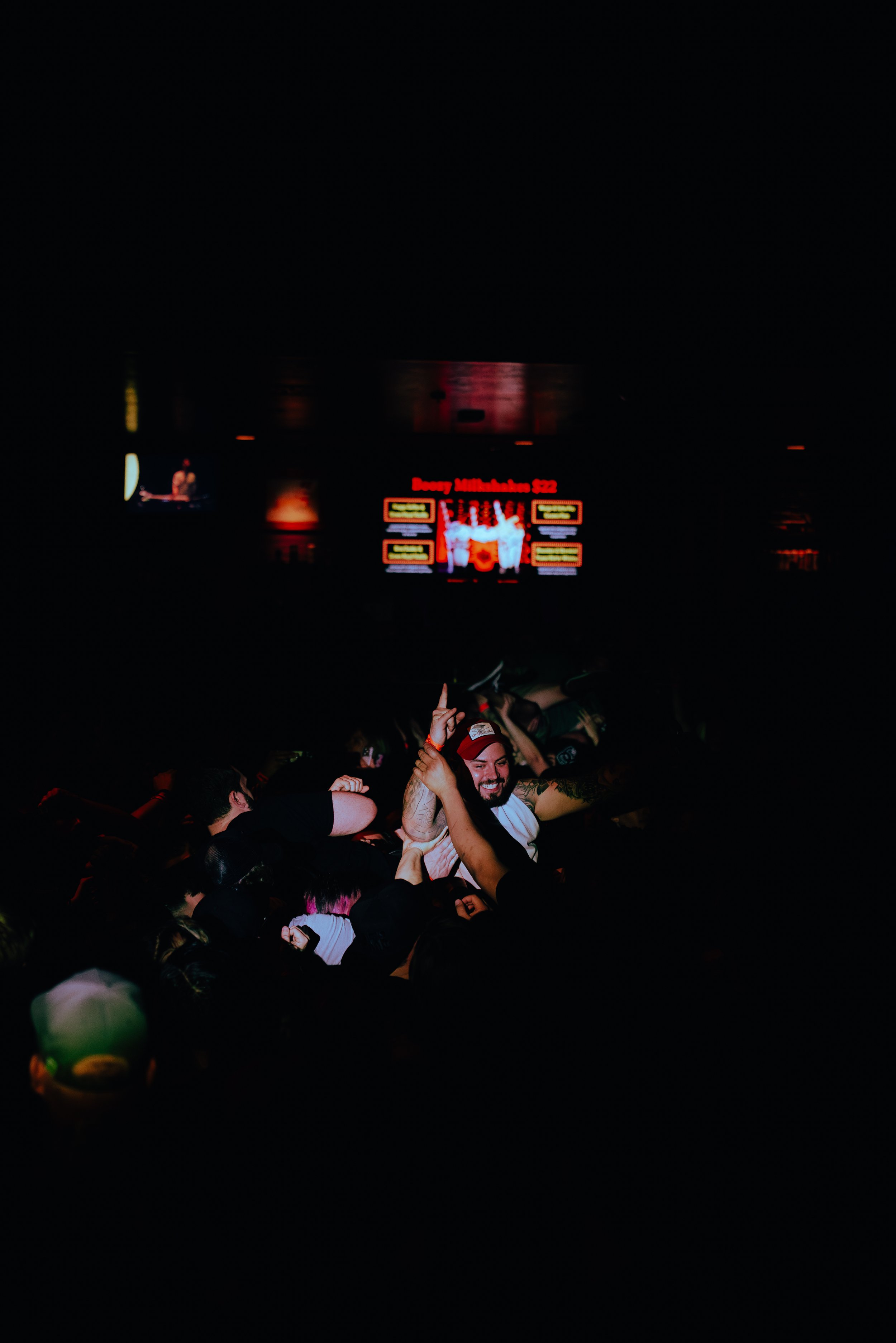 Crowd of people enjoying a concert or event at night, with a large screen displaying colorful graphics and red text in the background.