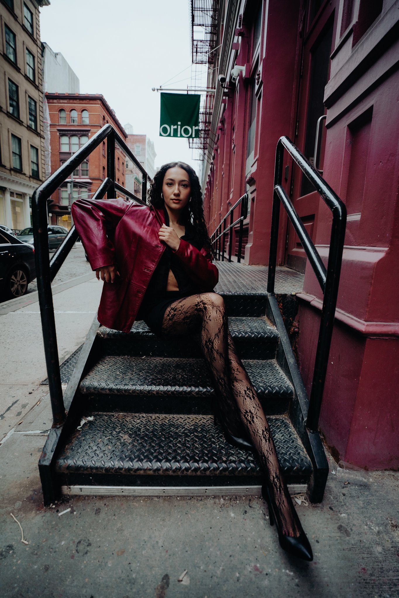 A woman with long dark curly hair is sitting on black metal stairs outside a red brick building. She is wearing a red leather jacket, fishnet stockings, and black boots. The background shows city buildings and a street.