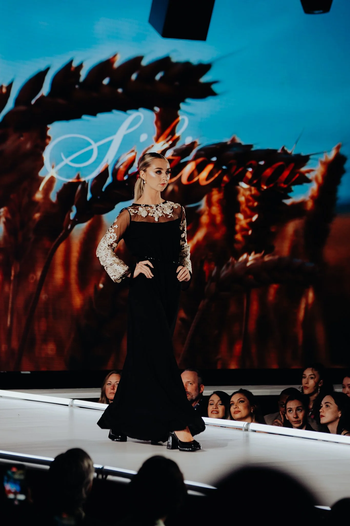A woman in a black dress with lace sleeves walks on runway during a fashion show, with audience members watching and a large screen in the background displaying colorful graphics and text.