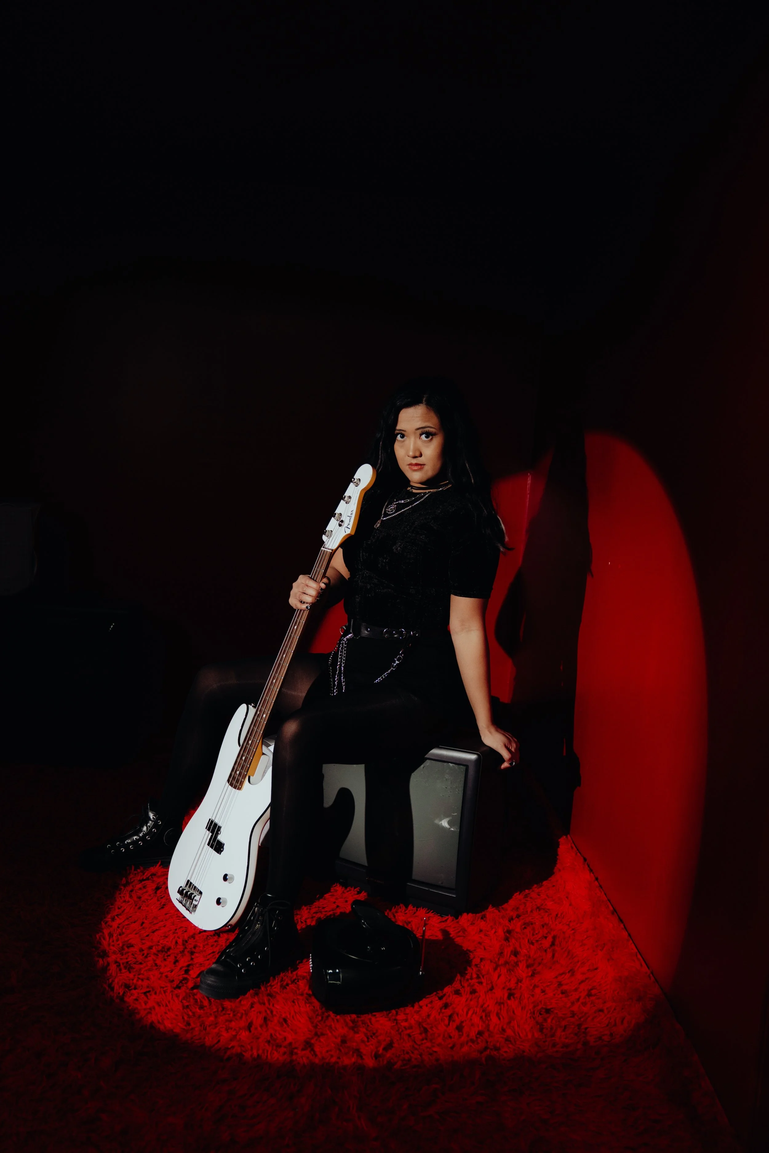 A woman with long dark hair and dressed in black, sitting on a chair with a white electric bass guitar. She is in a dark room with black walls, a red textured rug, and a red backdrop. She looks directly at the camera with a serious expression.