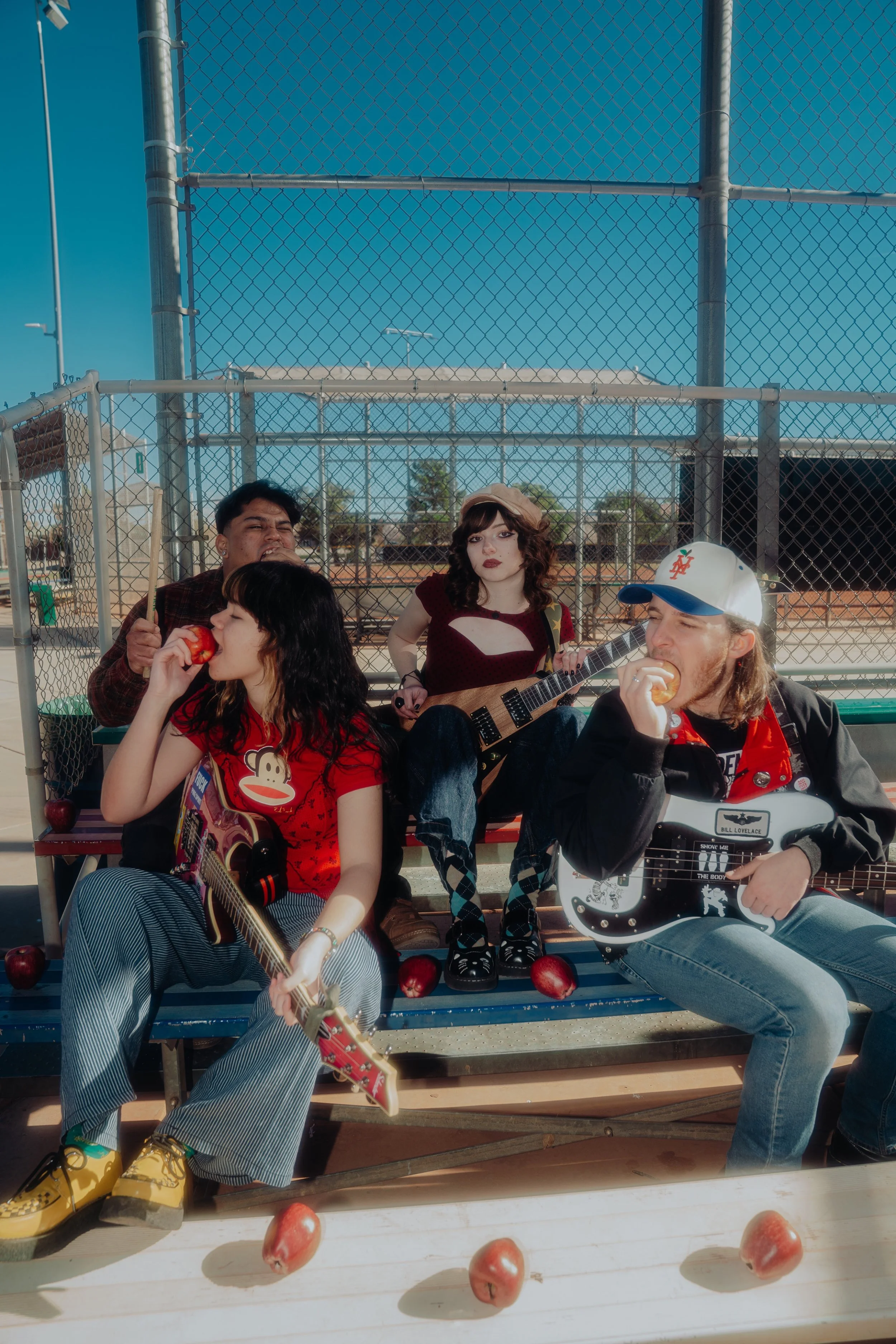 A group of young people sitting on bleachers at a baseball field, eating apples and playing guitars, with a chain-link fence and clear blue sky in the background.