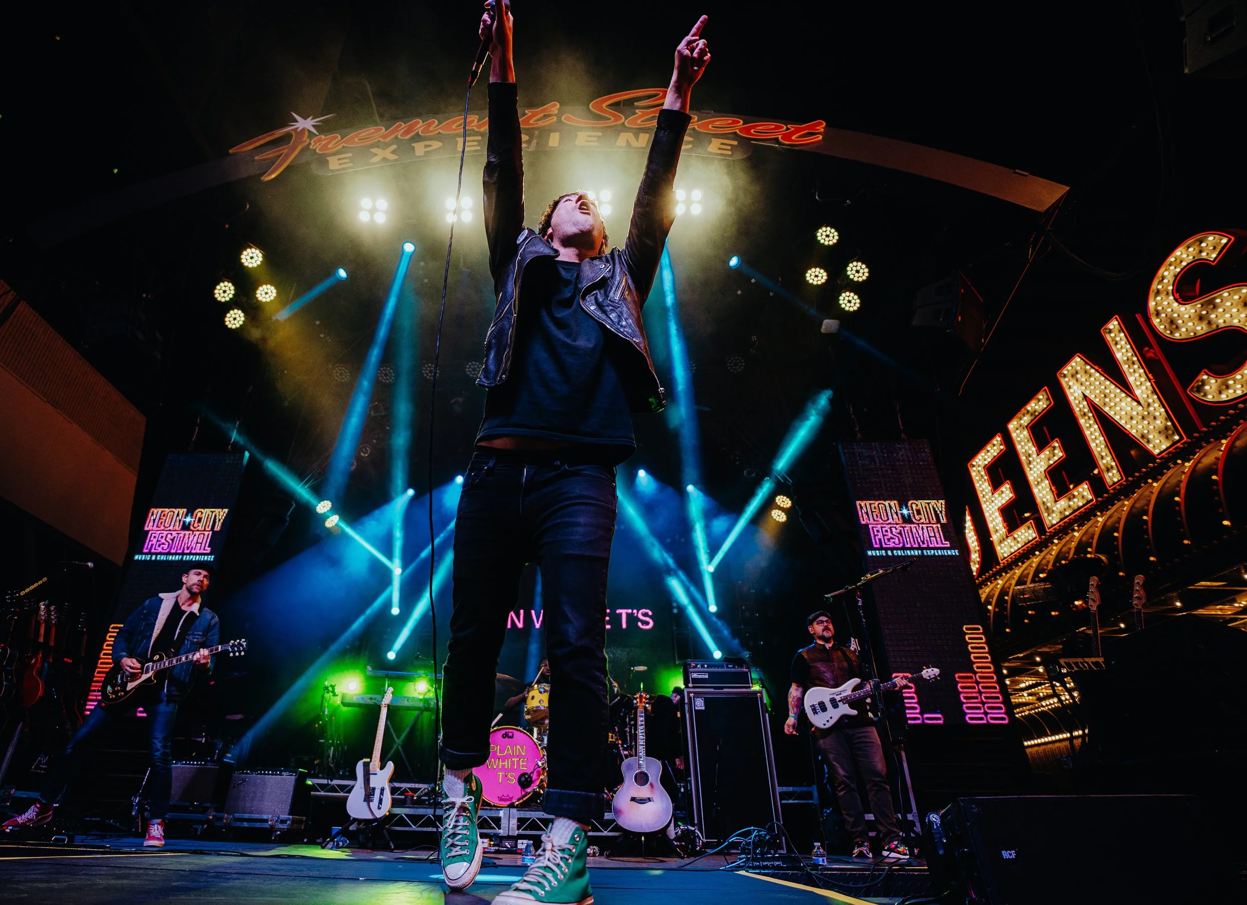 A band performing live on stage at the Neon City Festival, with bright colorful lights and signage in the background. The lead singer is in the center, raising one hand. Other band members are playing guitars, with one wearing green sneakers.
