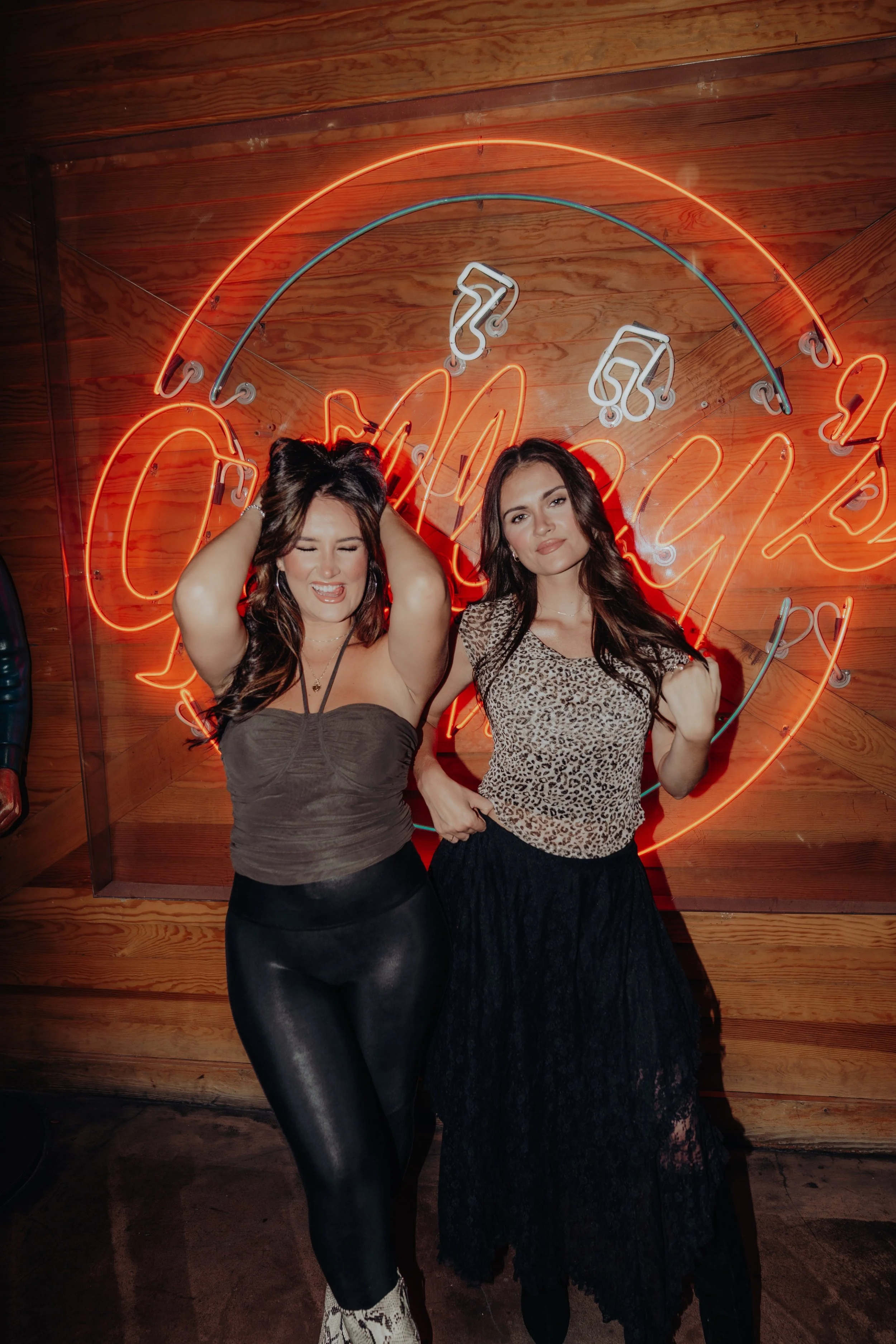 Two women standing in front of a neon sign with a wooden wall background. The woman on the left has long brown hair, is wearing a gray strapless top and black faux leather pants, with one arm raised and eyes closed. The woman on the right has long da