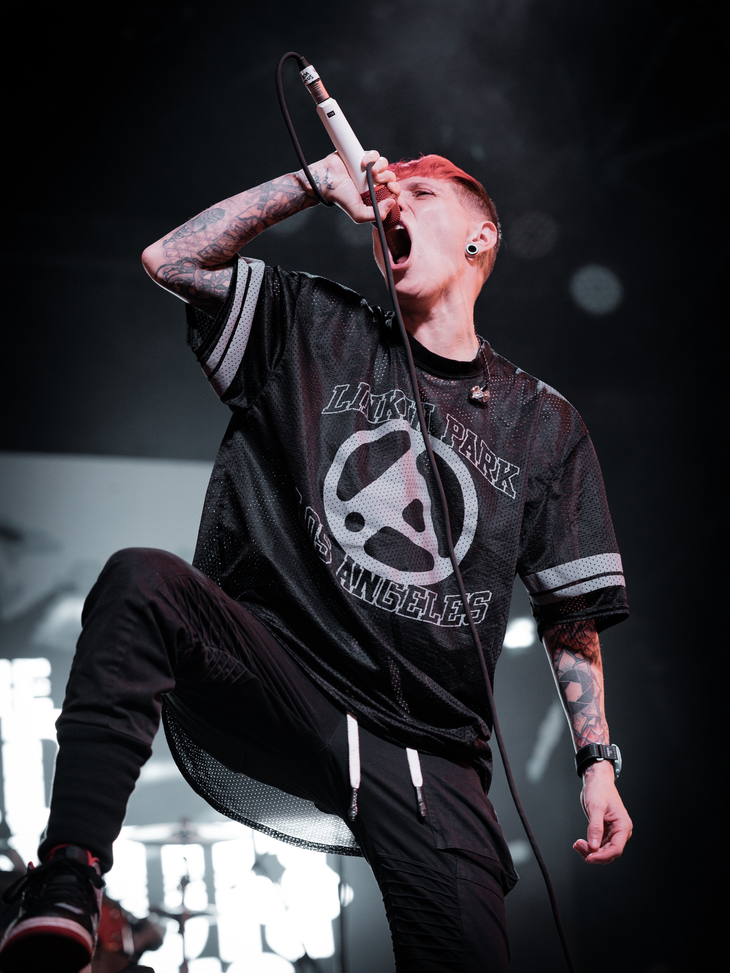 A vocalist with short red hair and tattoos on their arm sings passionately into a microphone on stage, wearing a black and gray band shirt and black pants.