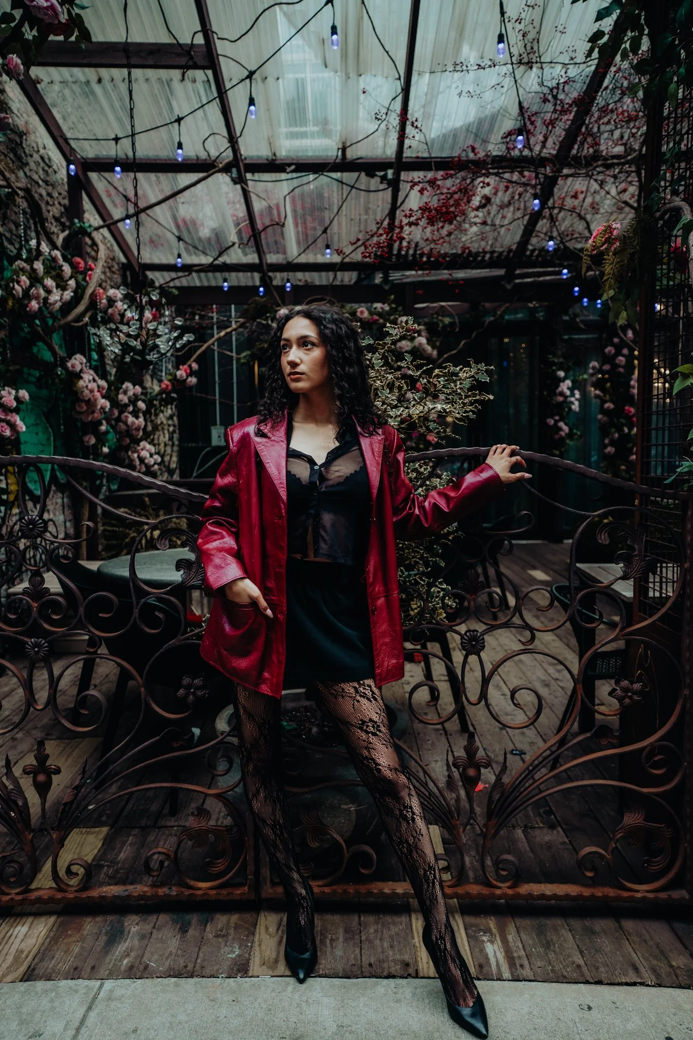 A woman with curly dark hair wearing a red leather jacket over a black mesh top, black lace stockings, and black heels, standing on a decorative wrought iron fence in a lush garden with pink flowers and hanging string lights.