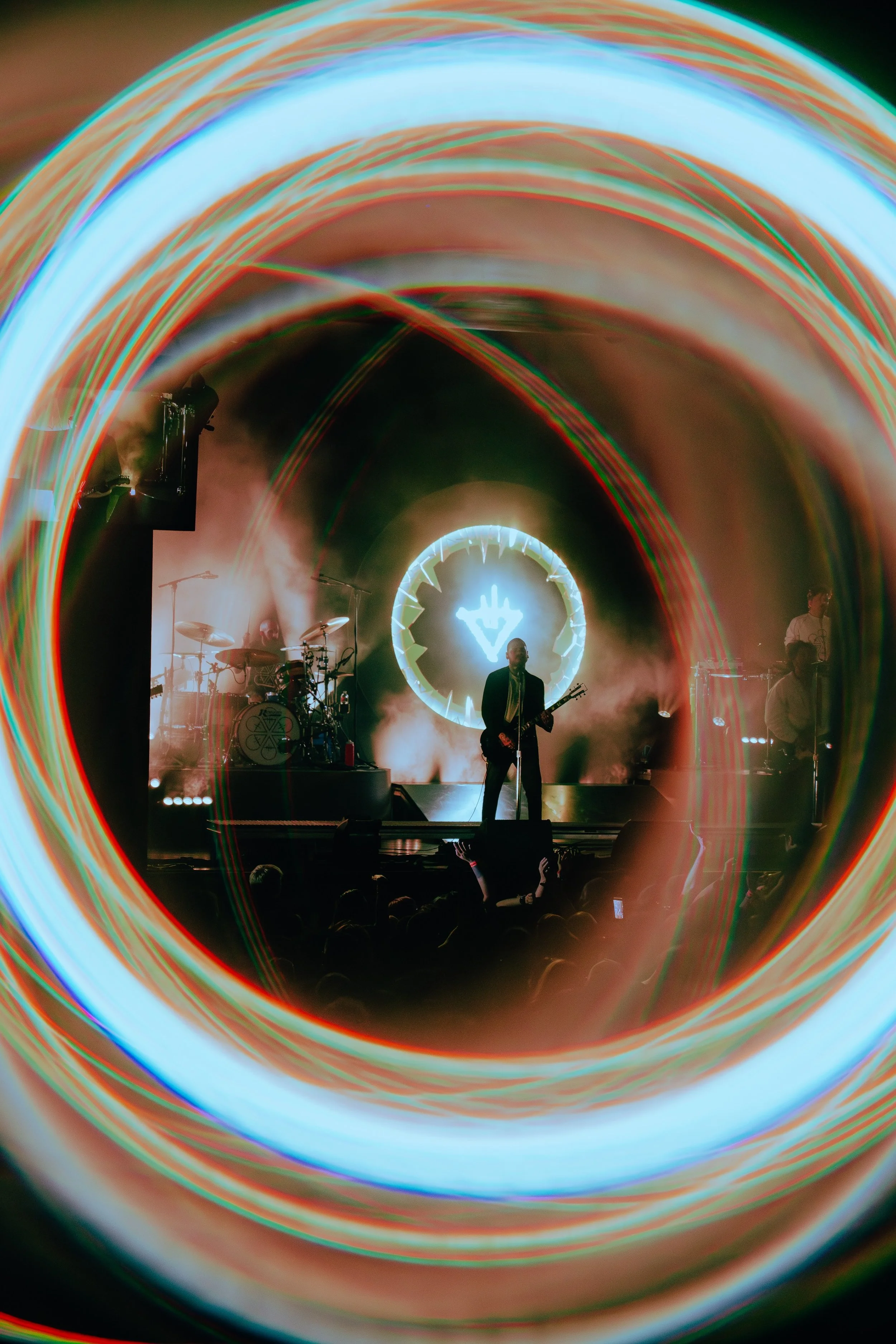 A live music performance on stage with colorful light trails creating a tunnel effect around the performers. The band is playing, with one musician in the center holding a guitar, and a drummer and other band members are visible in the background. Br