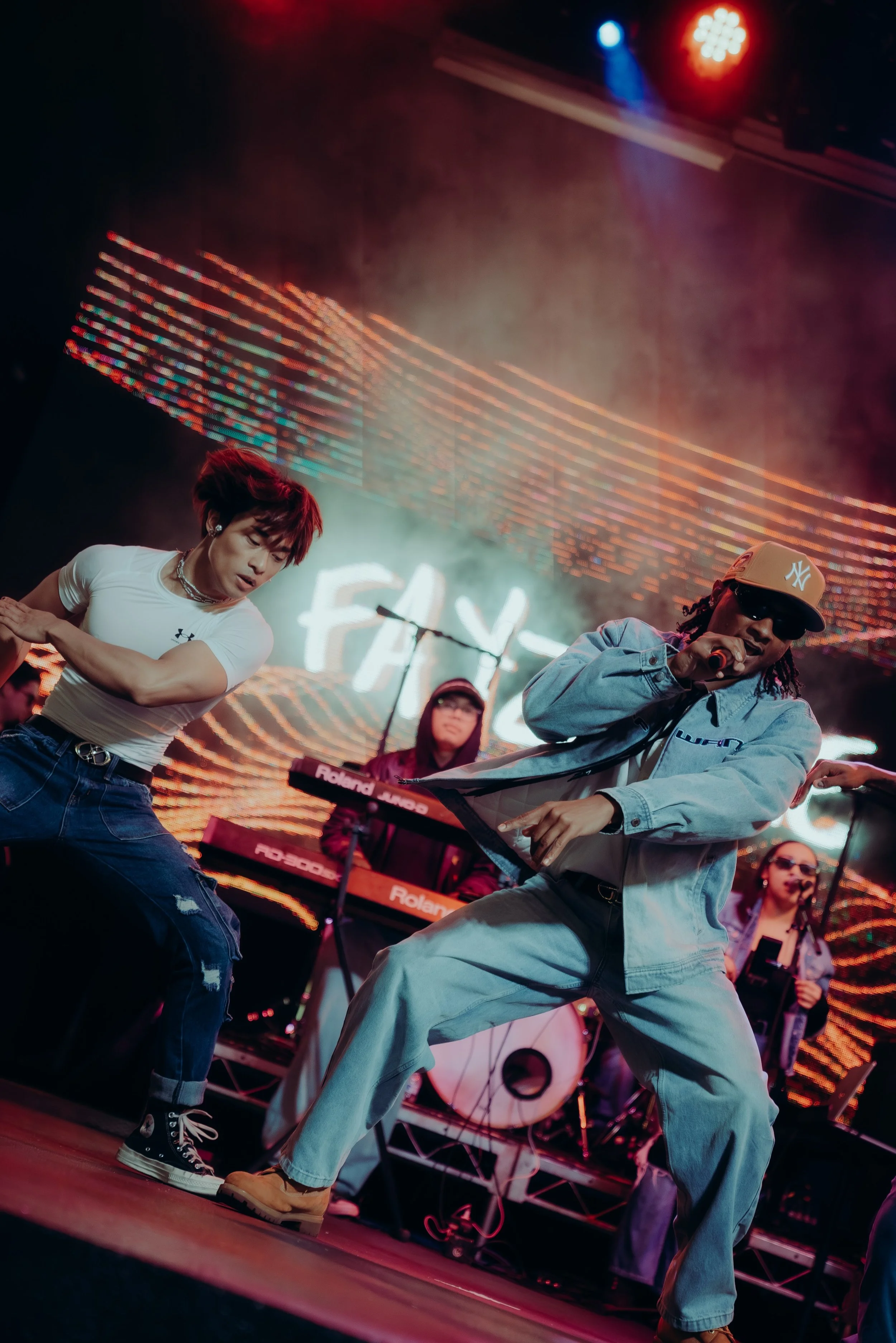 A group of performers dancing passionately on stage with colorful lighting and electronic music equipment in the background.