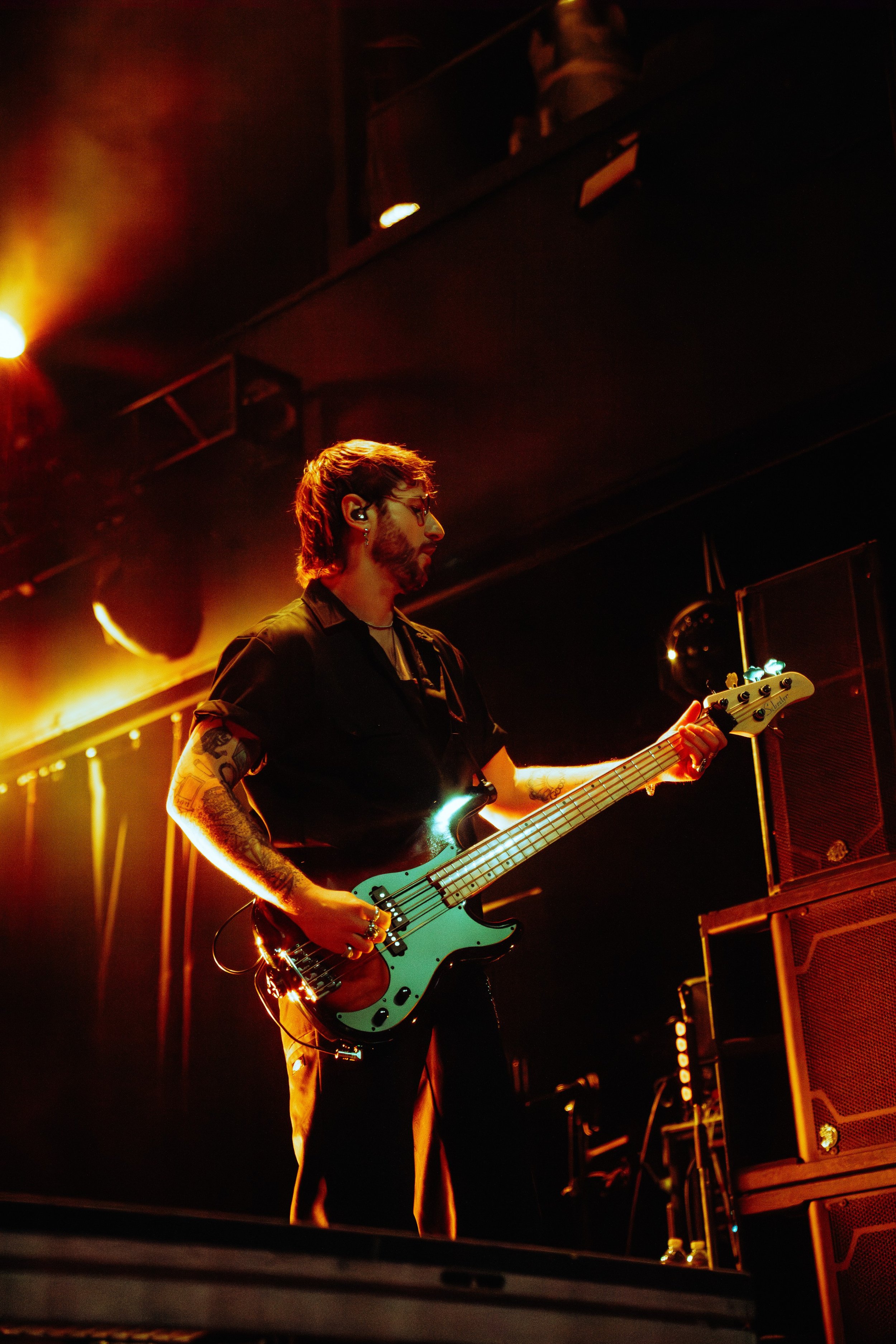 A musician with tattoos and glasses playing an electric bass guitar on stage with yellow stage lighting and amplifiers in the background.