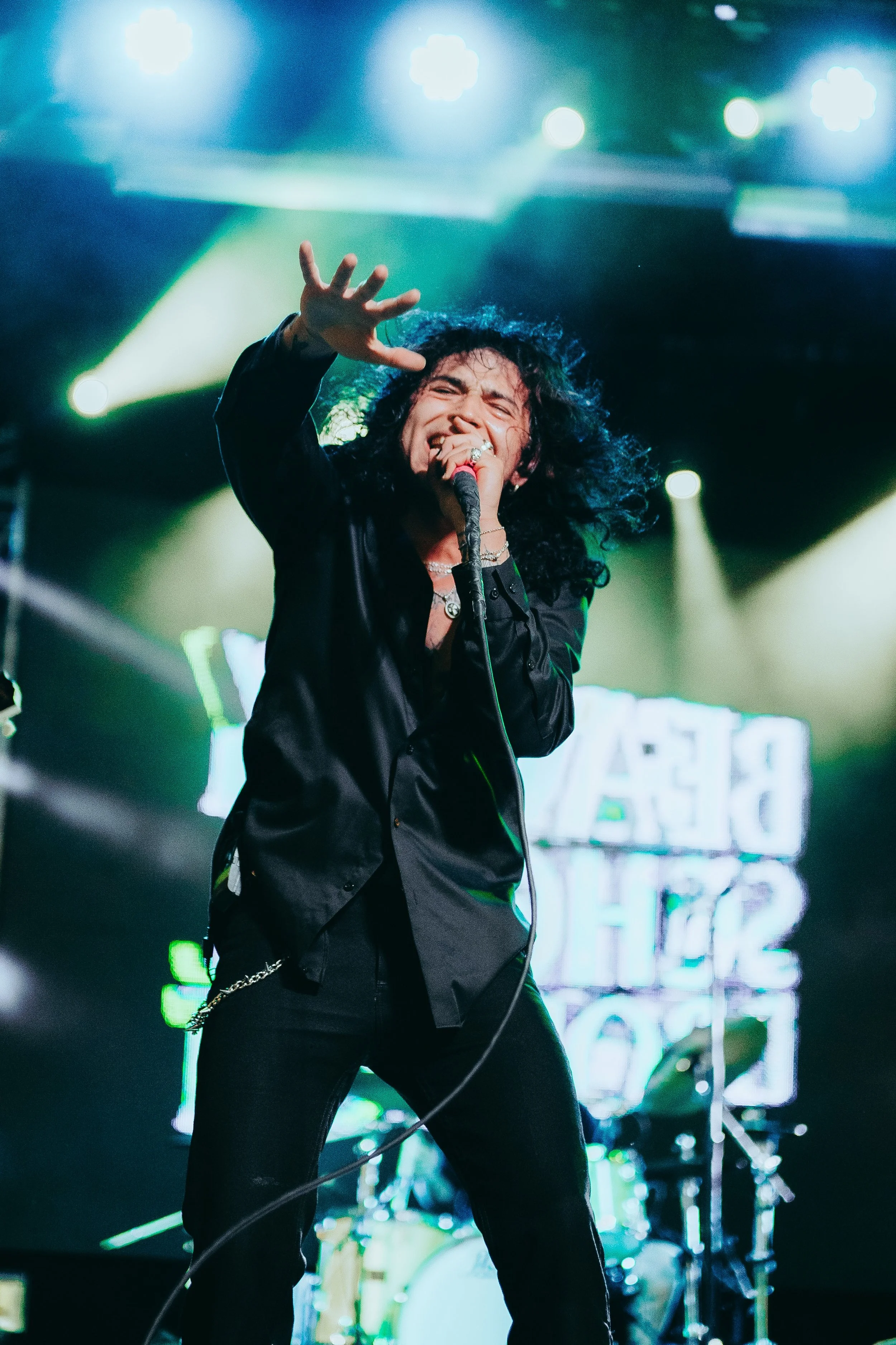 A male vocalist with long curly hair passionately singing into a microphone on stage, with stage lights and a drum set in the background.