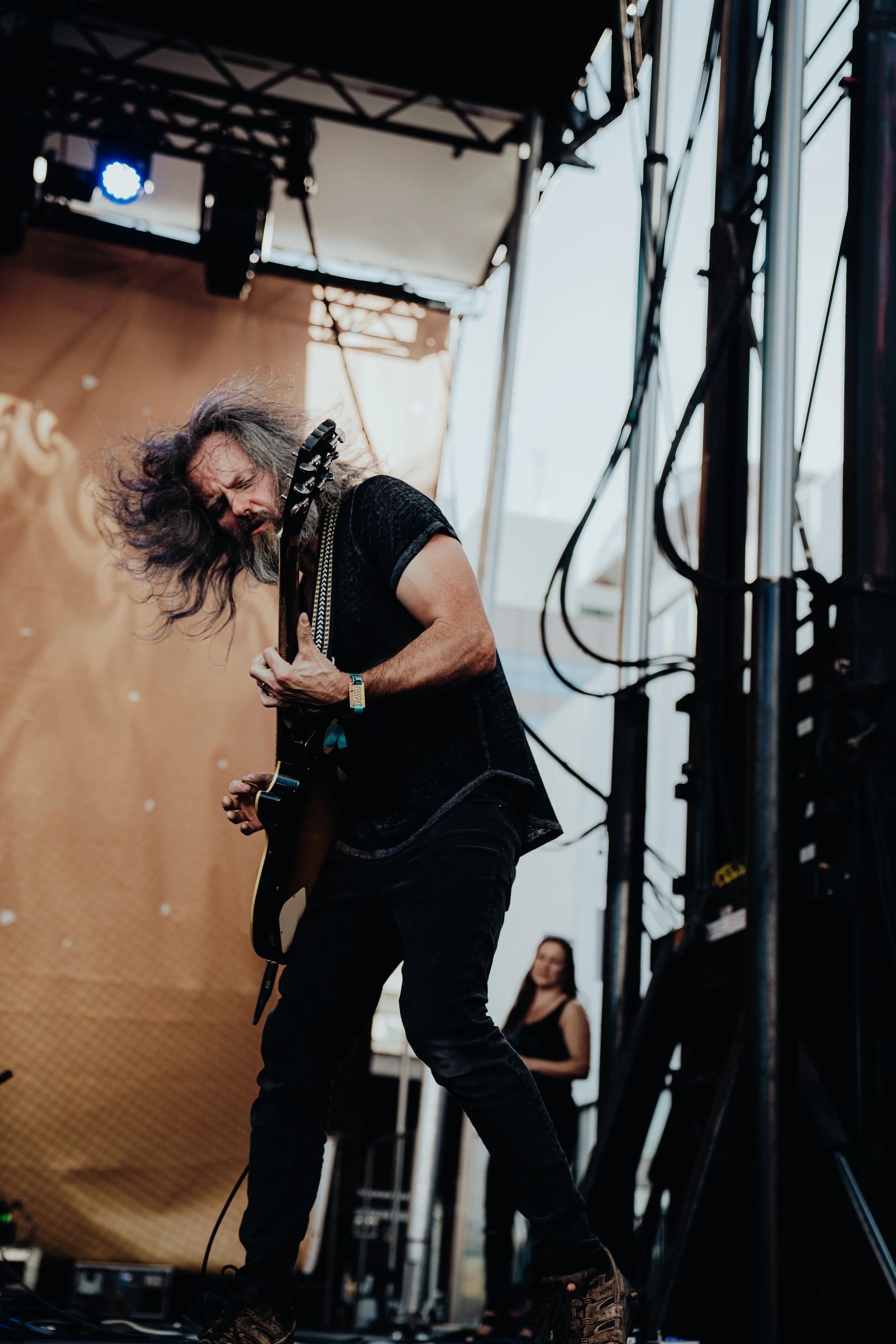 A male guitarist with long hair and beard playing electric guitar on stage during a performance, with stage equipment and a woman in the background.