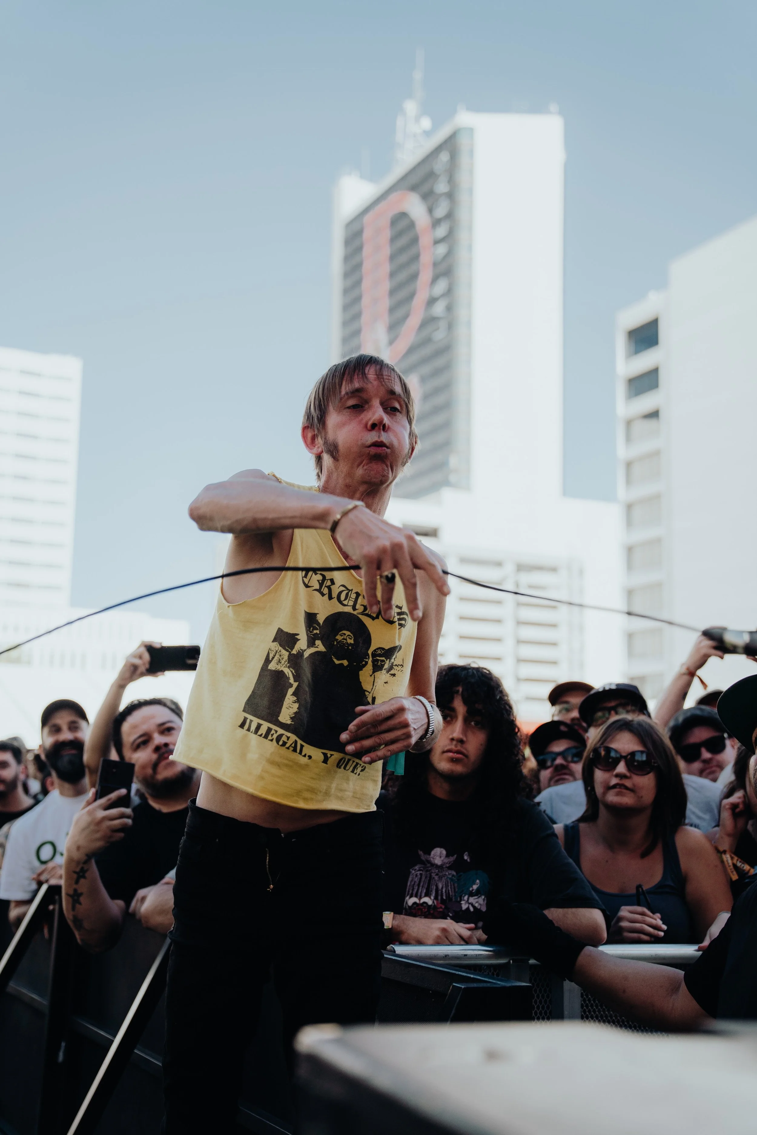 A man in a yellow T-shirt with a graphic design and the words 'Illegal, Y O' standing in front of a crowd, during a protest or rally, with tall buildings in the background, including one with a large illuminated heart symbol.