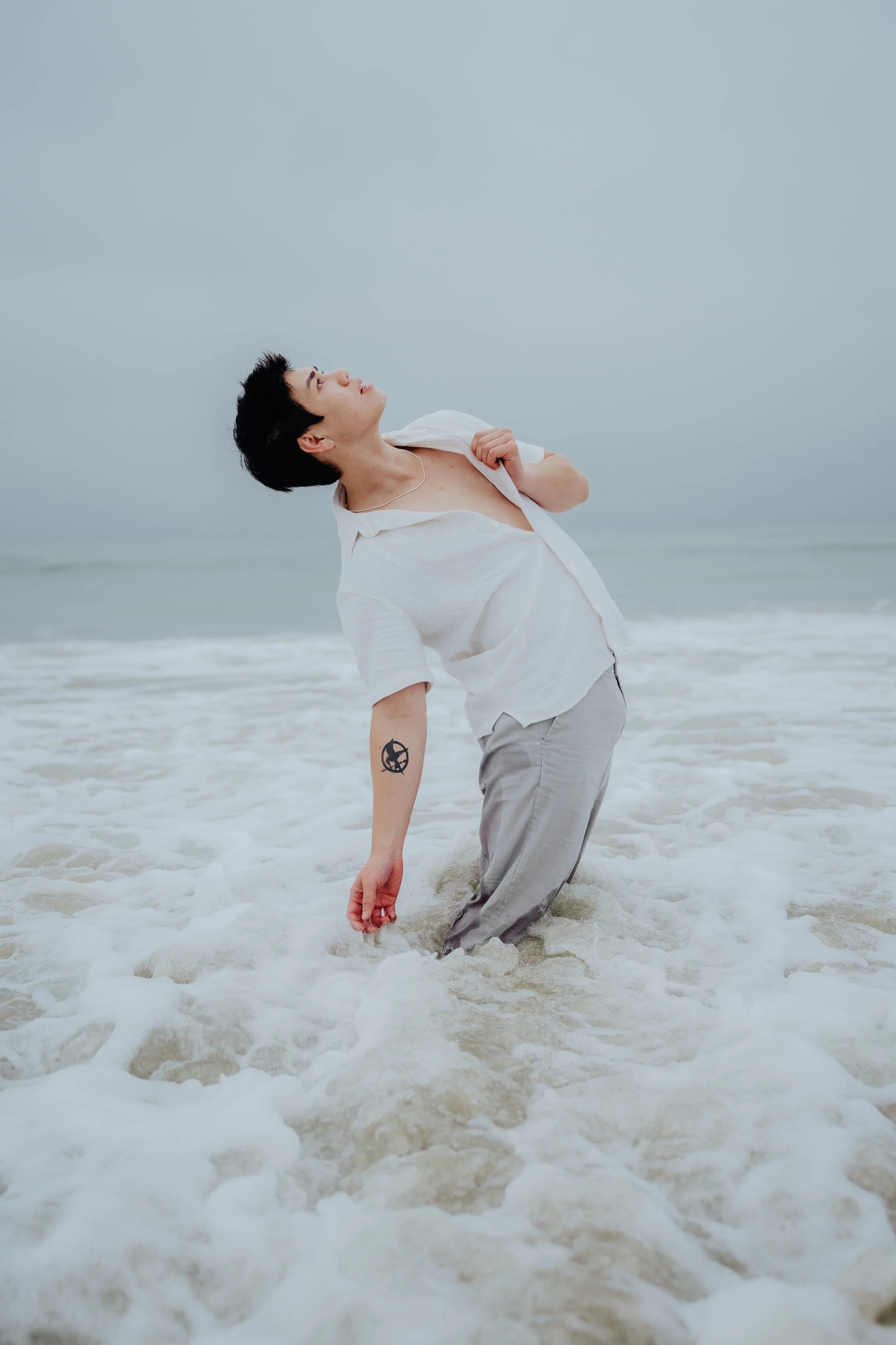 A young man with short black hair and a tattoo on his left forearm stands in shallow ocean water, leaning back with water up to his knees, as he gazes upward against a cloudy sky. He wears a white open shirt and light-colored pants.
