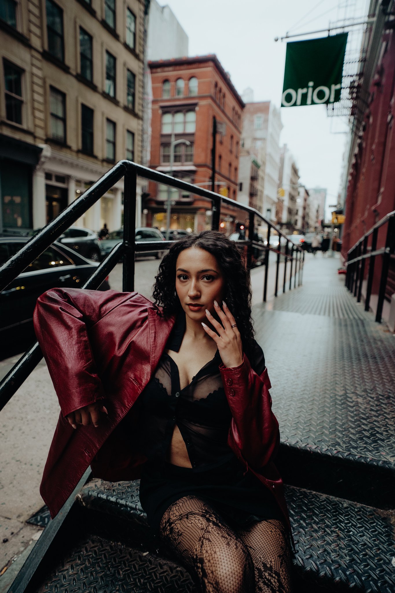 A young woman with curly black hair and light skin seated outdoors on a metal platform, wearing black fishnet stockings, a black lace top, and a red leather jacket draped over her shoulder, with a city street scene in the background.
