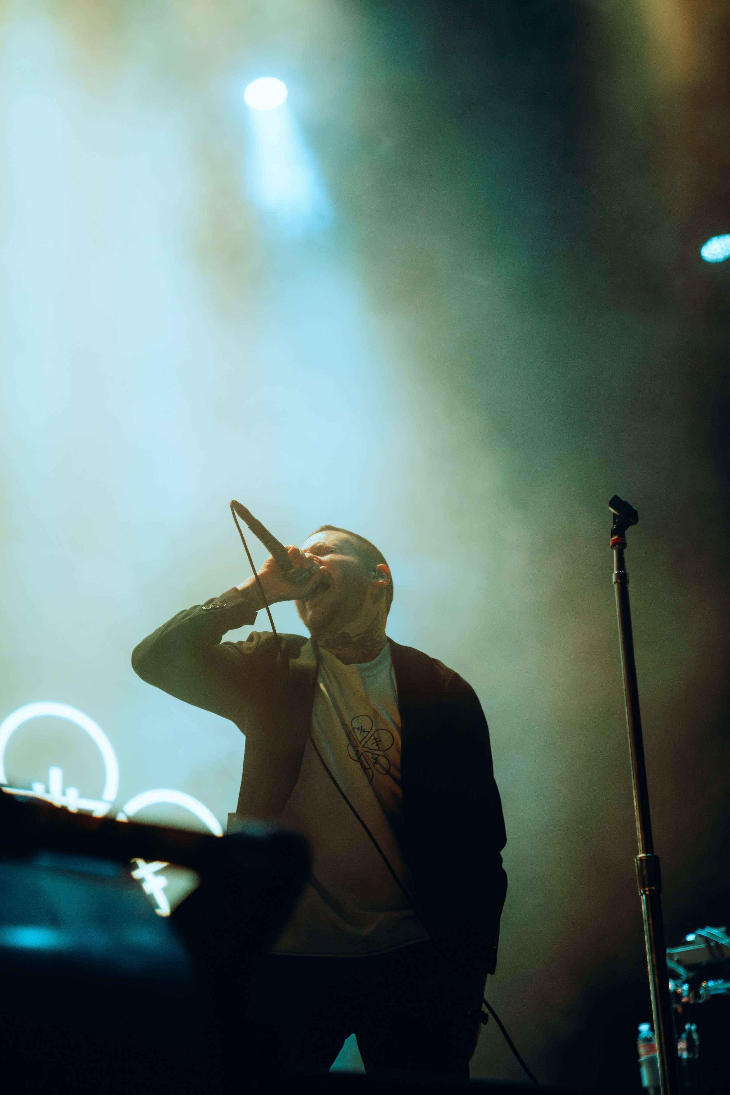 A man singing into a microphone on stage with stage lights and fog in the background.