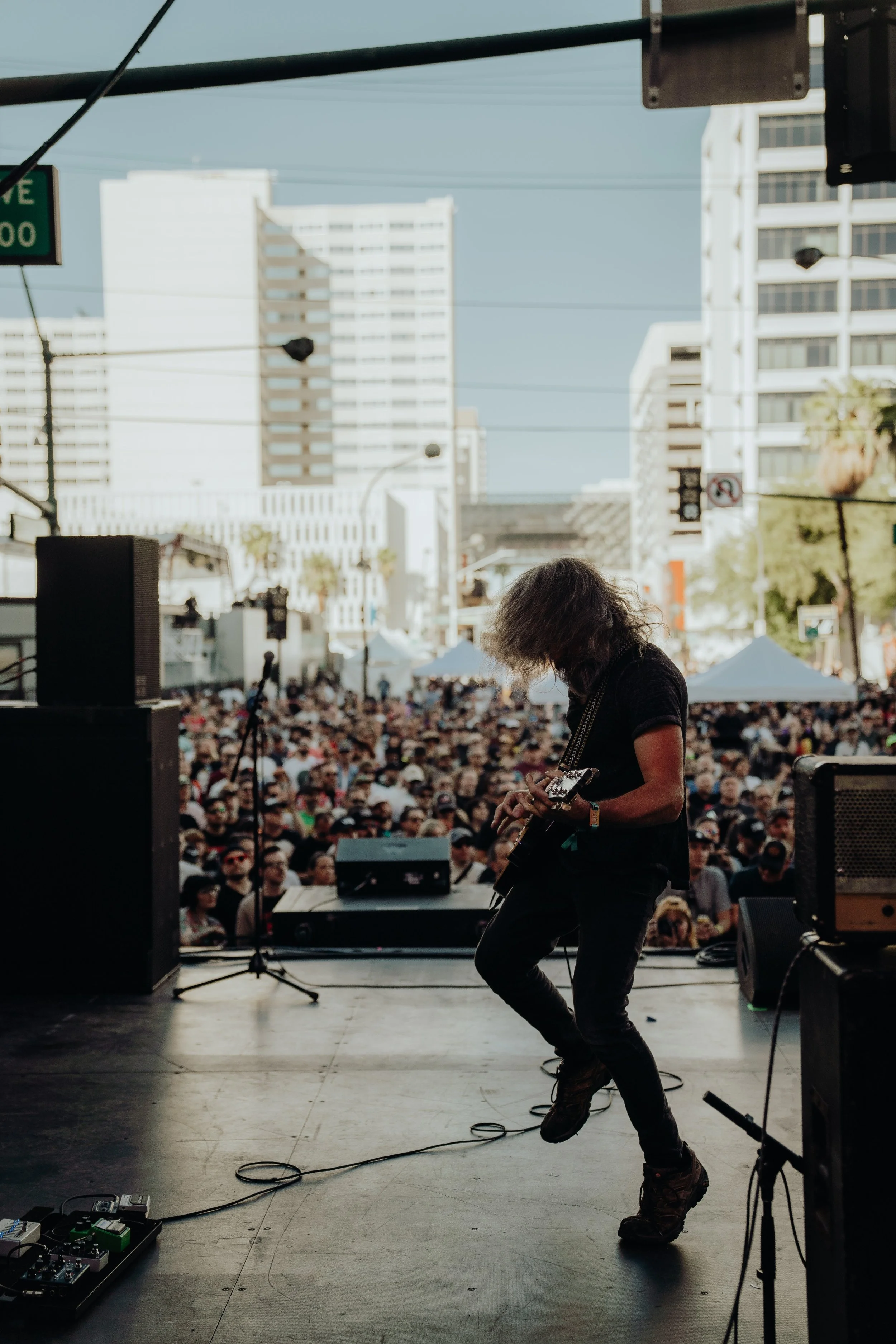A musician playing an electric guitar on stage with a large crowd in front and tall buildings in the background during an outdoor event.