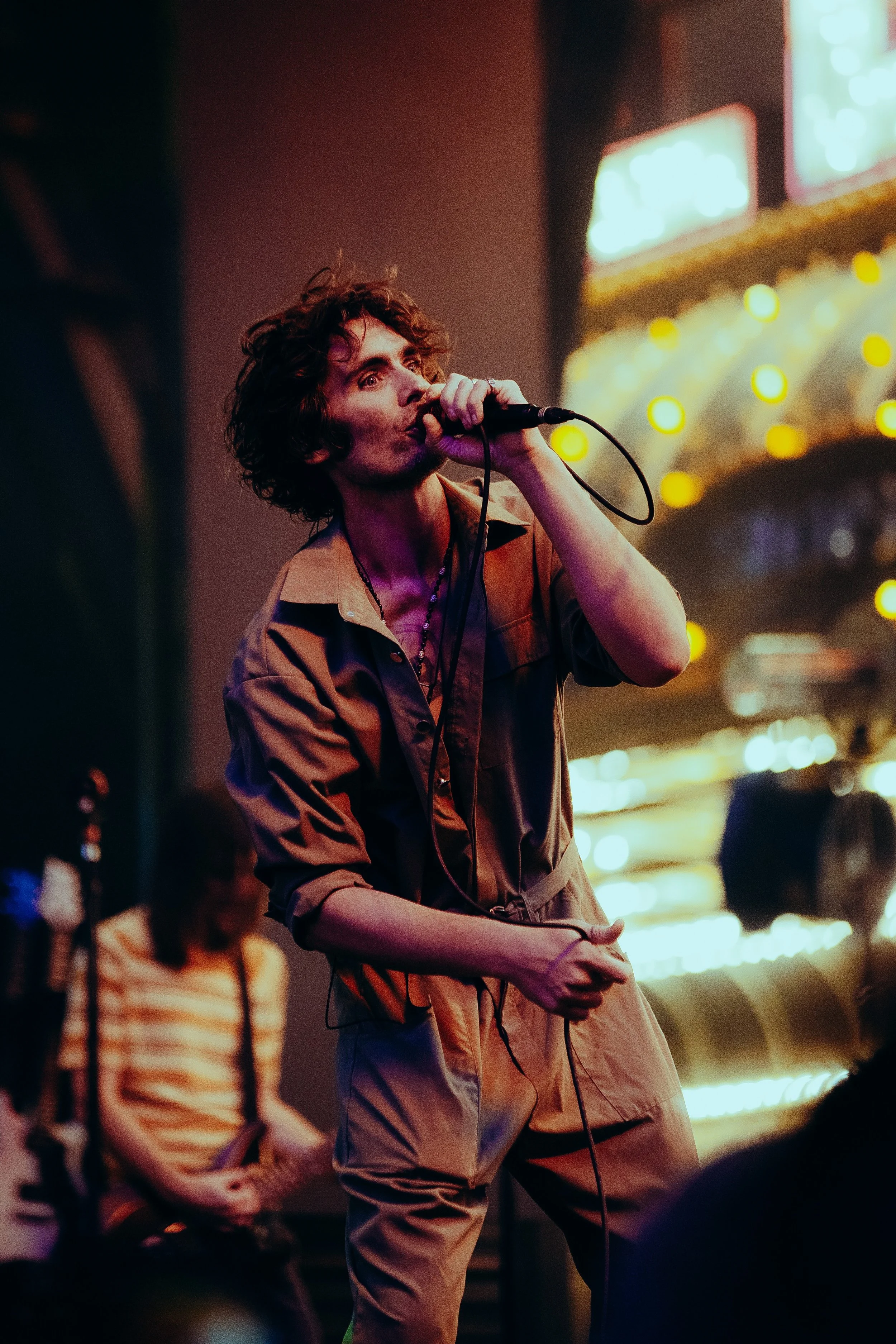 A person with curly hair singing into a microphone in front of a blurred background with bright lights and signs.