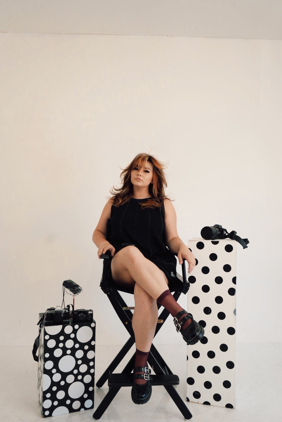 A woman with reddish-brown hair, wearing a black sleeveless top and black shoes, sitting on a black folding chair in front of a plain light-colored wall. She has one leg crossed over the other, and her expression is neutral. To her left and right, there are black-and-white polka dot boxes with cameras on top, and a camera on a strap on the right box.