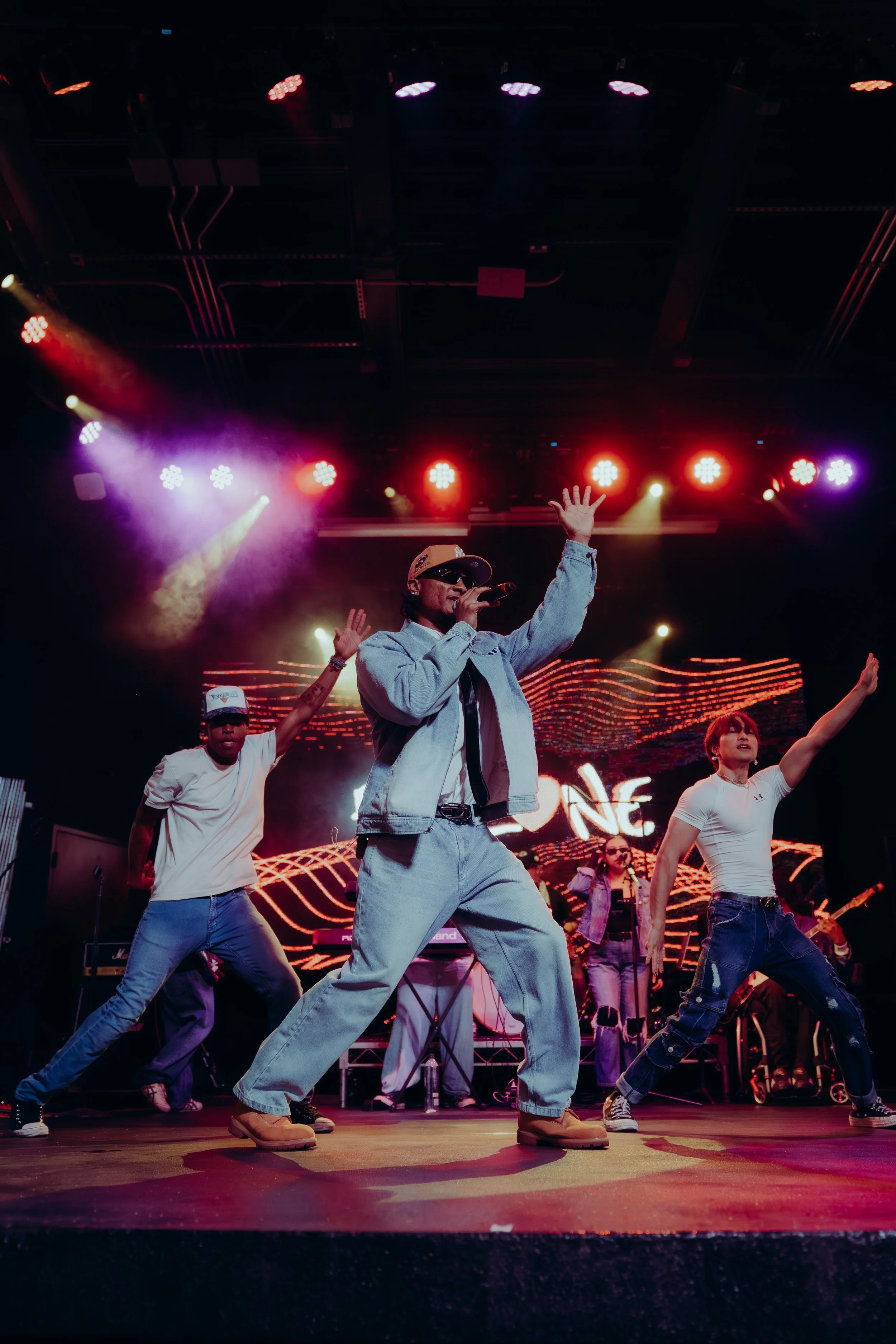 Performer singing and dancing on stage with colorful lights, wearing sunglasses, denim jacket, and beige boots, with backup dancers in casual clothes and a digital background.