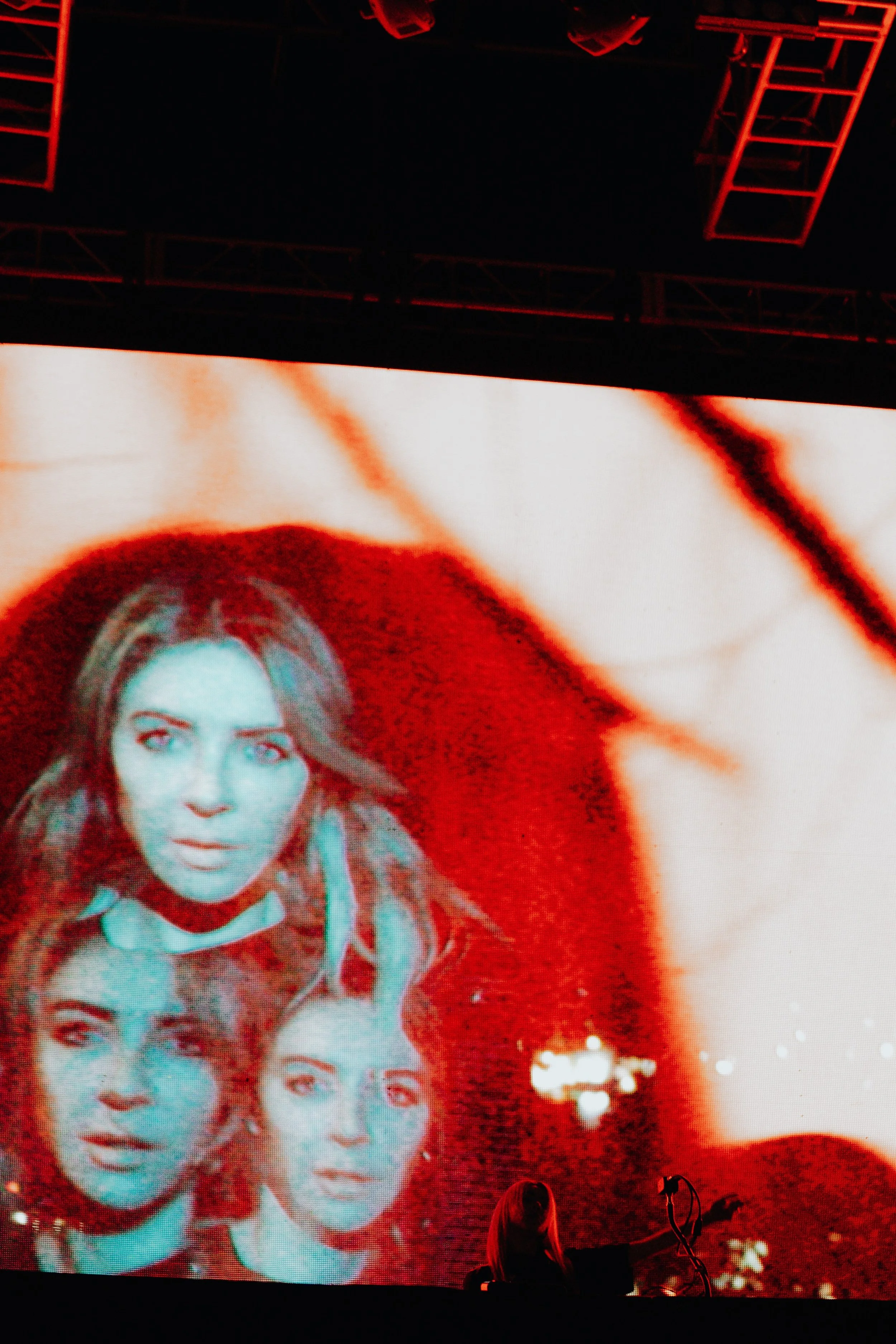 Large screen displaying a stylized image of a woman with long hair and serious expression, rendered in red tones. The scene is set in a dark room with stage lighting, and a person is visible seated at a piano in the lower part of the image.