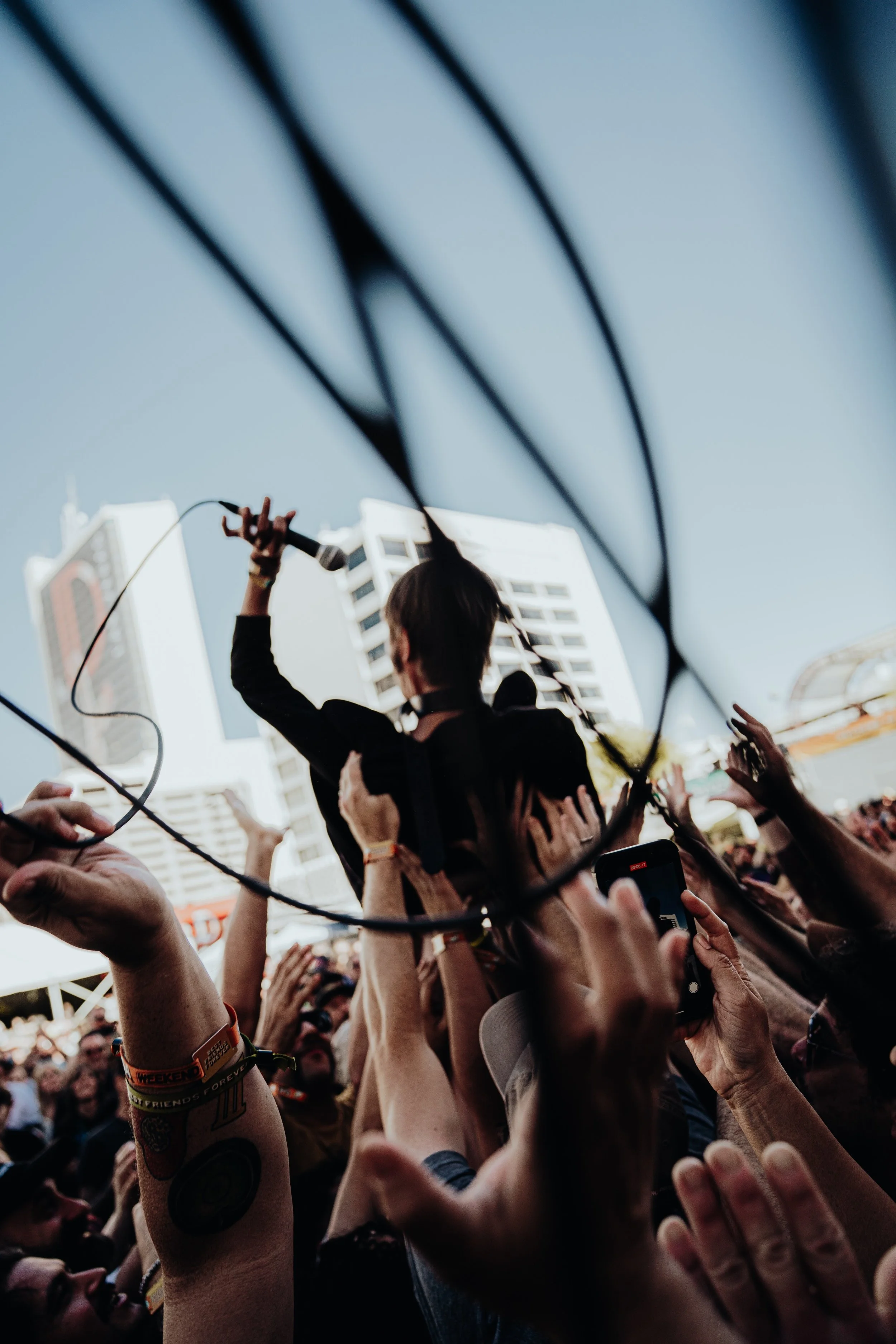 Crowd of people raising hands at an outdoor event with a person on stage holding a microphone. Tall buildings are visible in the background.