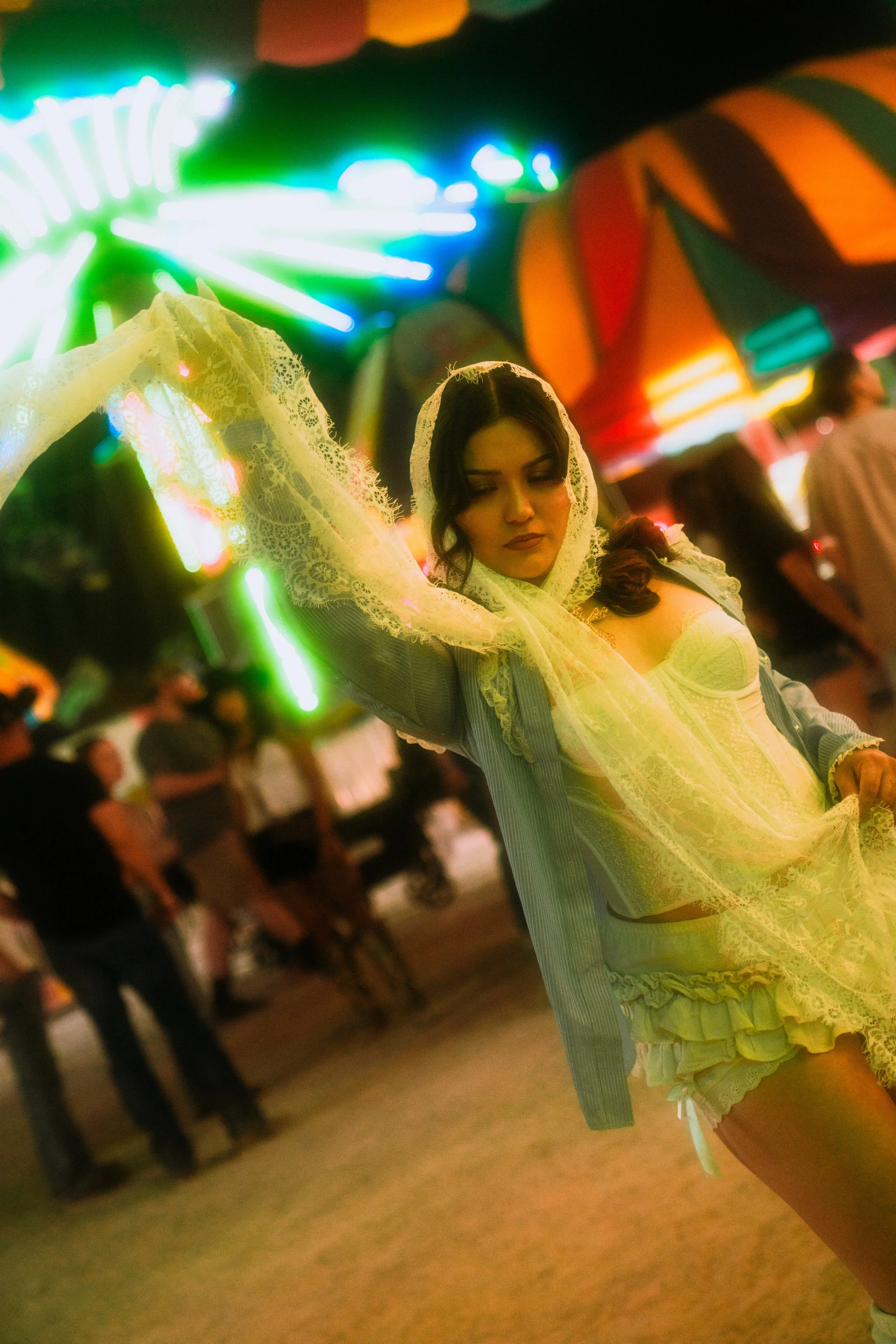A woman in a vintage dress with lace details and a veil is dancing at a fair or carnival at night, with a blurred Ferris wheel with colorful lights and other people in the background.