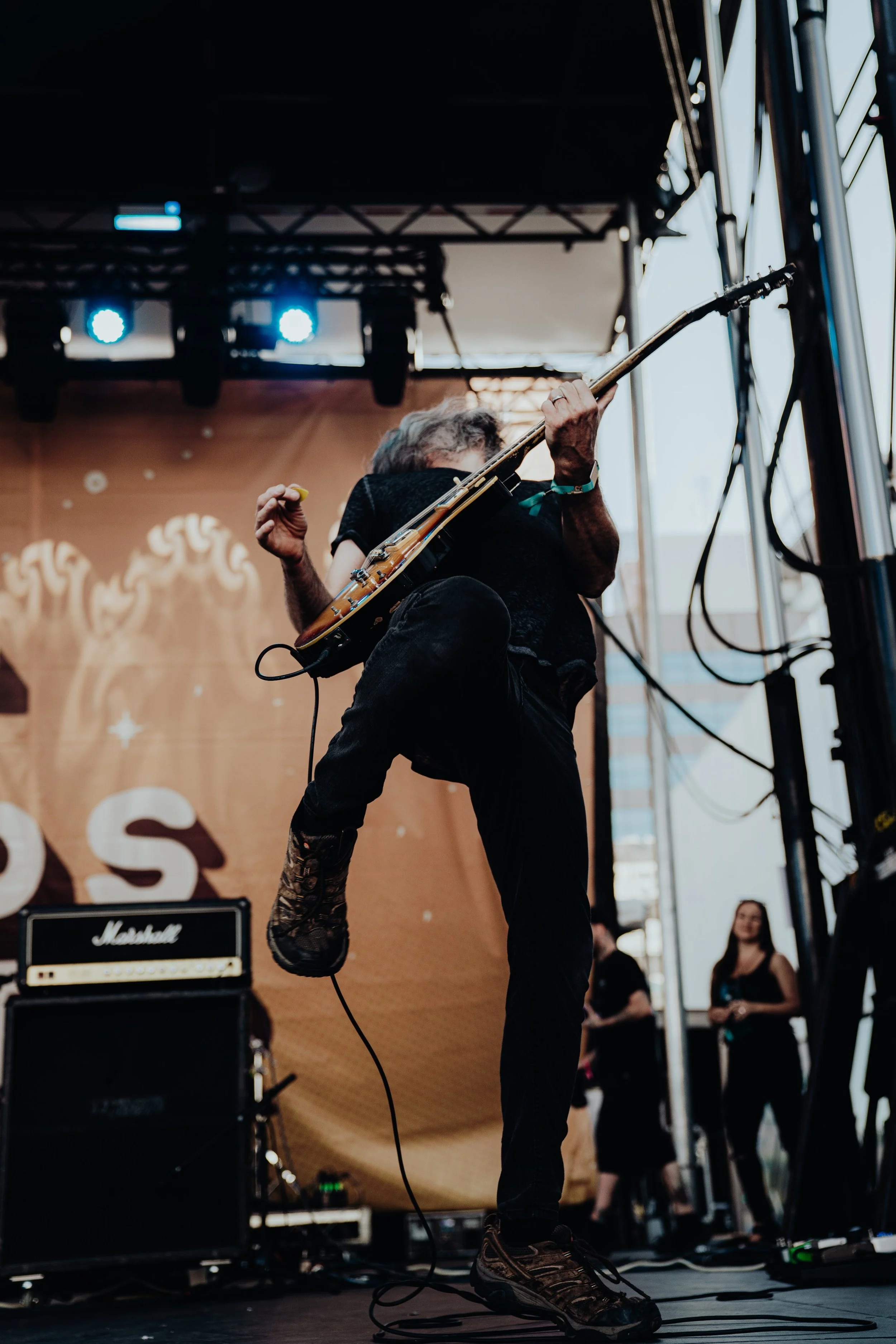 A musician performing on stage with a guitar, head bowed, one foot up on an amplifier, wearing black clothing and sneakers, with people watching in the background.