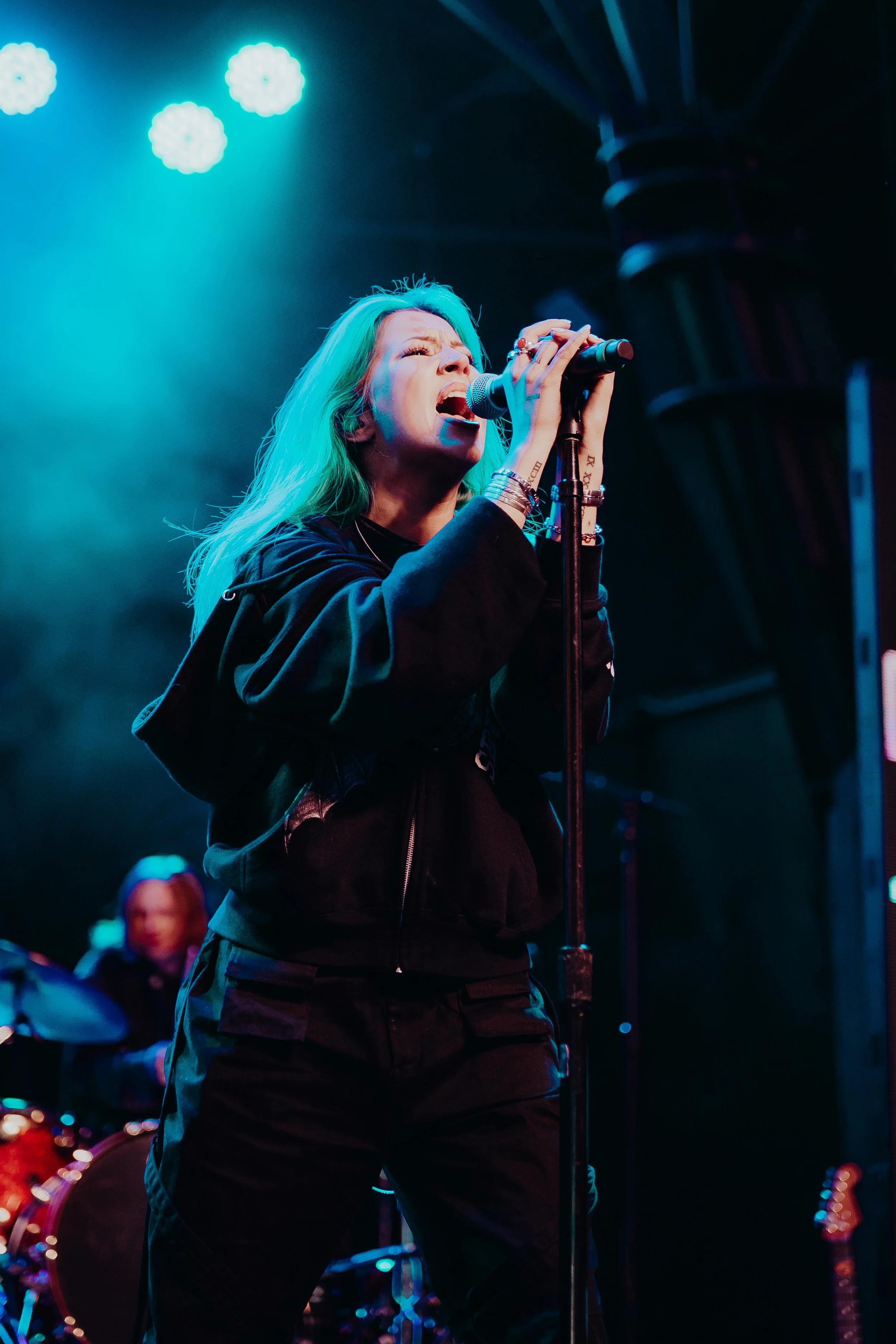 Woman singing passionately into a microphone on stage with stage lights overhead and a band in the background.