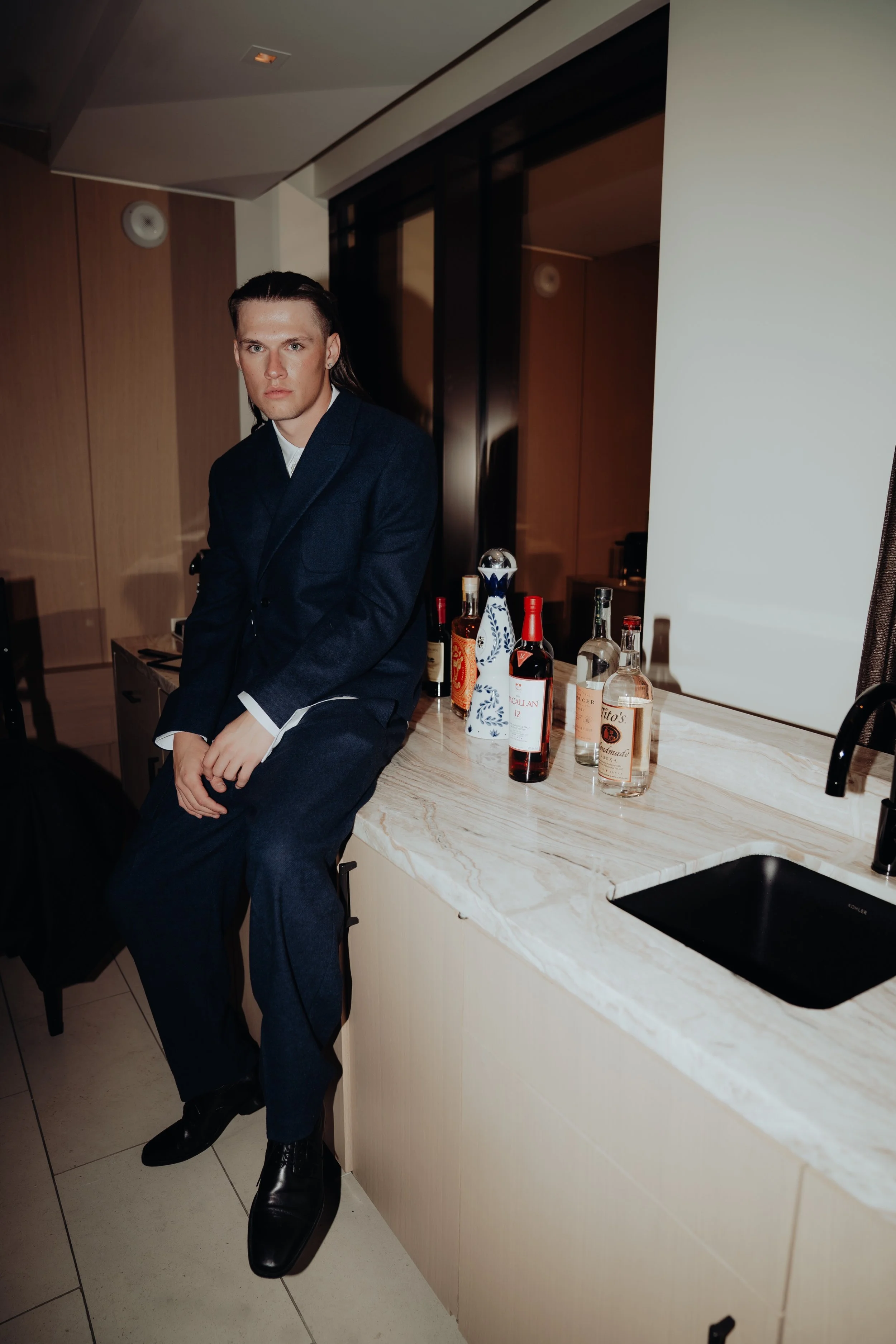 Man with long hair wearing a dark suit sitting on a kitchen counter with bottles of alcohol behind him.