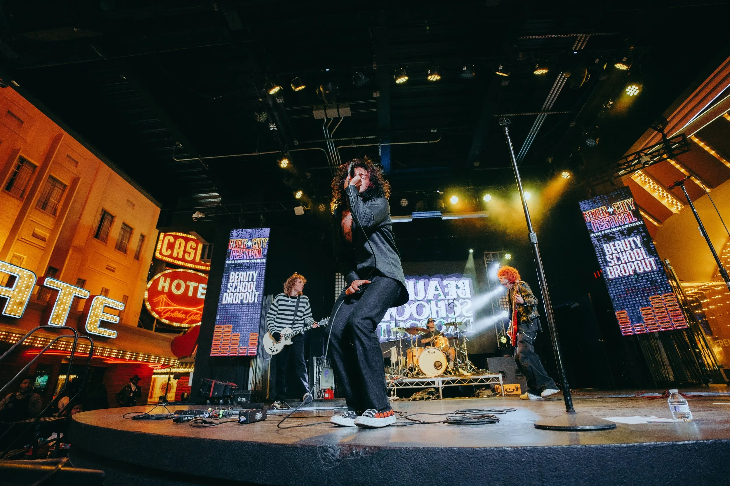 A band performs on an outdoor stage at night during a festival. The stage has large digital screens with colorful text, and setting with bright lights, neon signs, and surrounding buildings with illuminated signs.