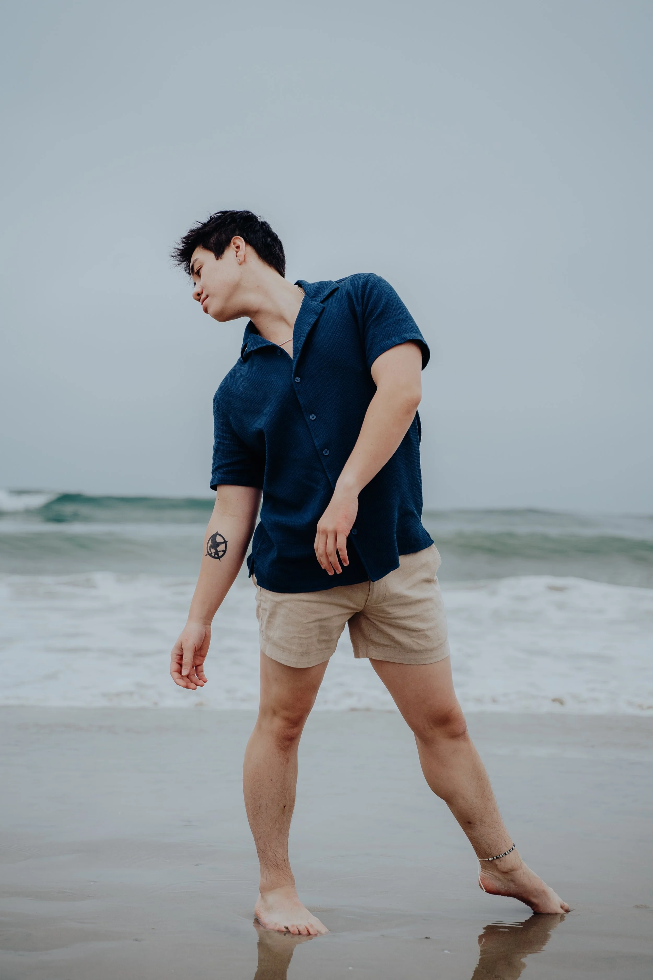 A young person with short dark hair, wearing a dark blue short-sleeved button-up shirt and beige shorts, standing on the beach with feet in the water, looking down, with a tattoo of a pentagram on their left forearm, against a cloudy sky and ocean wa