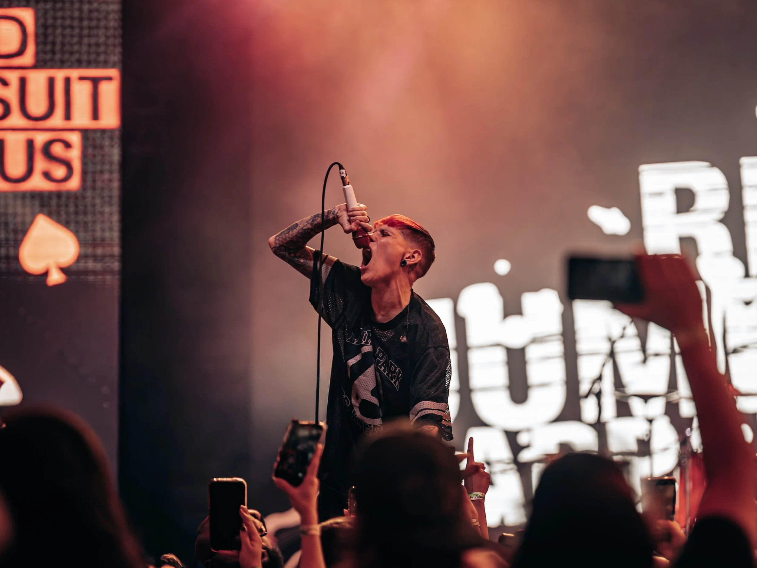 A person singing into a microphone during a live concert, with a crowd in front and neon signs in the background.