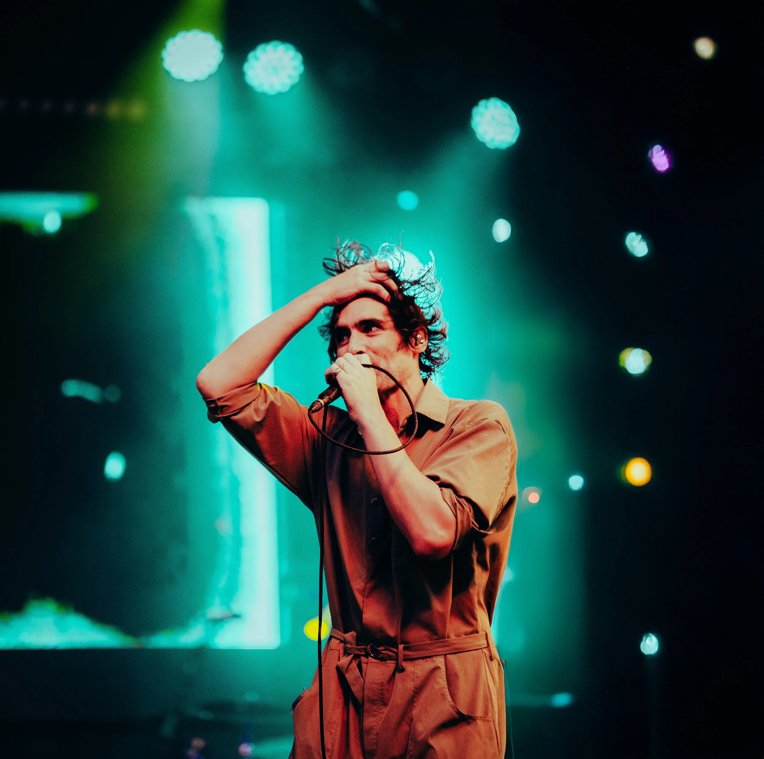 A person with curly hair holding a microphone and standing on stage with colorful lights in the background.