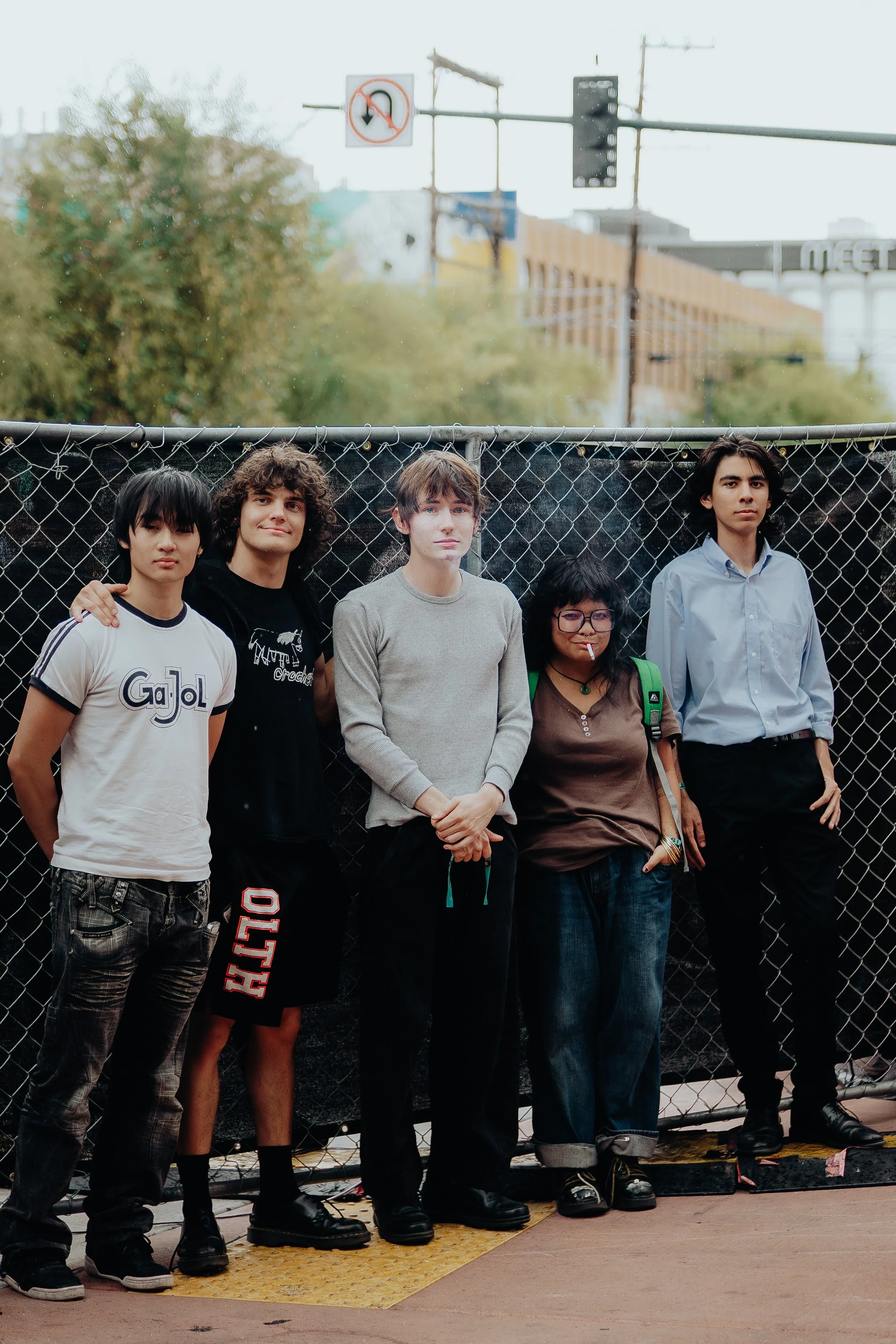 Five young people standing in front of a chain-link fence on an urban street, with trees and a building in the background. They are posing and looking at the camera.