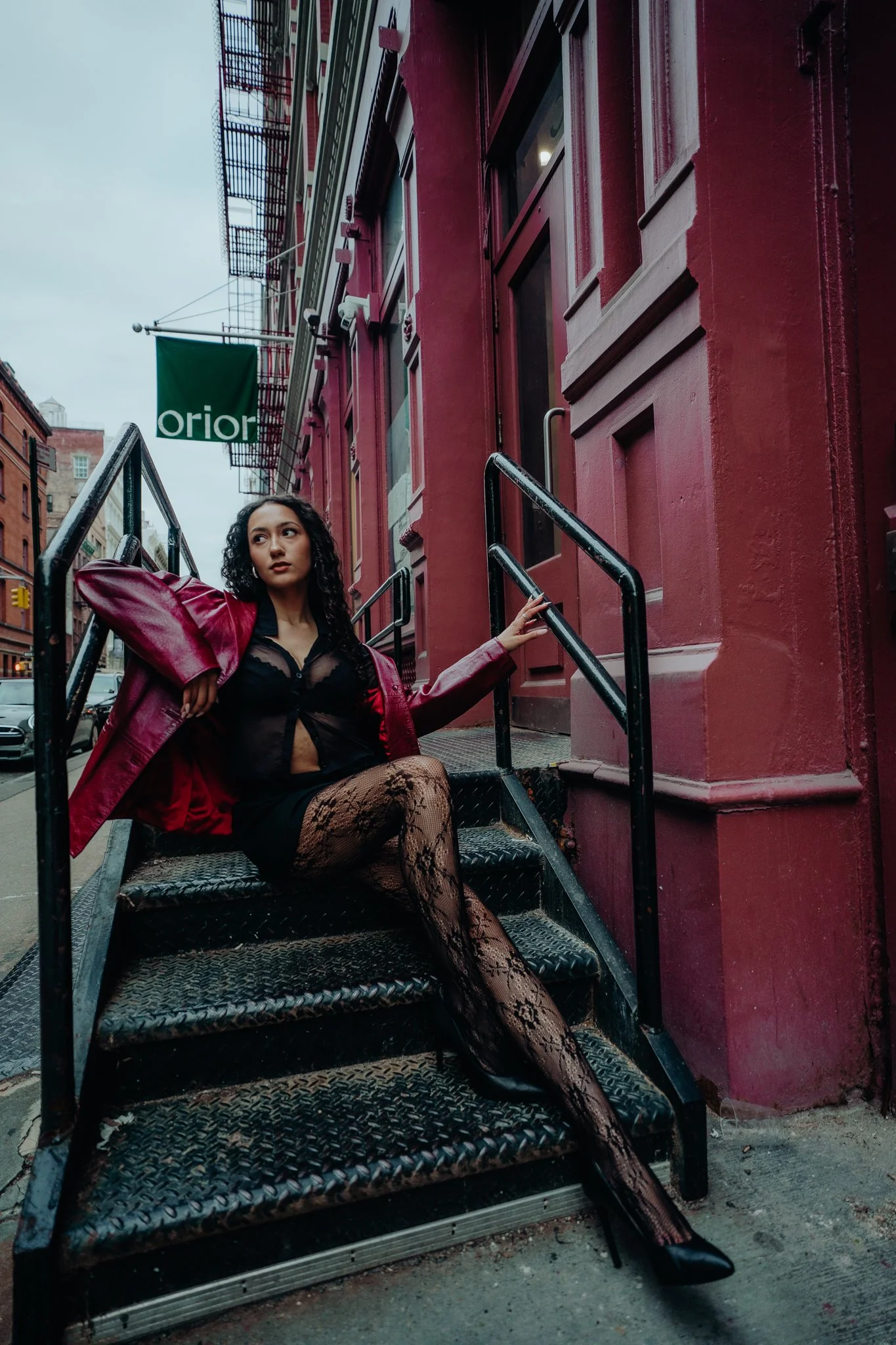 A woman with curly hair wearing a black top, lace stockings, and a red leather jacket sitting on metal stairs outside a red building with large windows and a green sign, under cloudy skies.