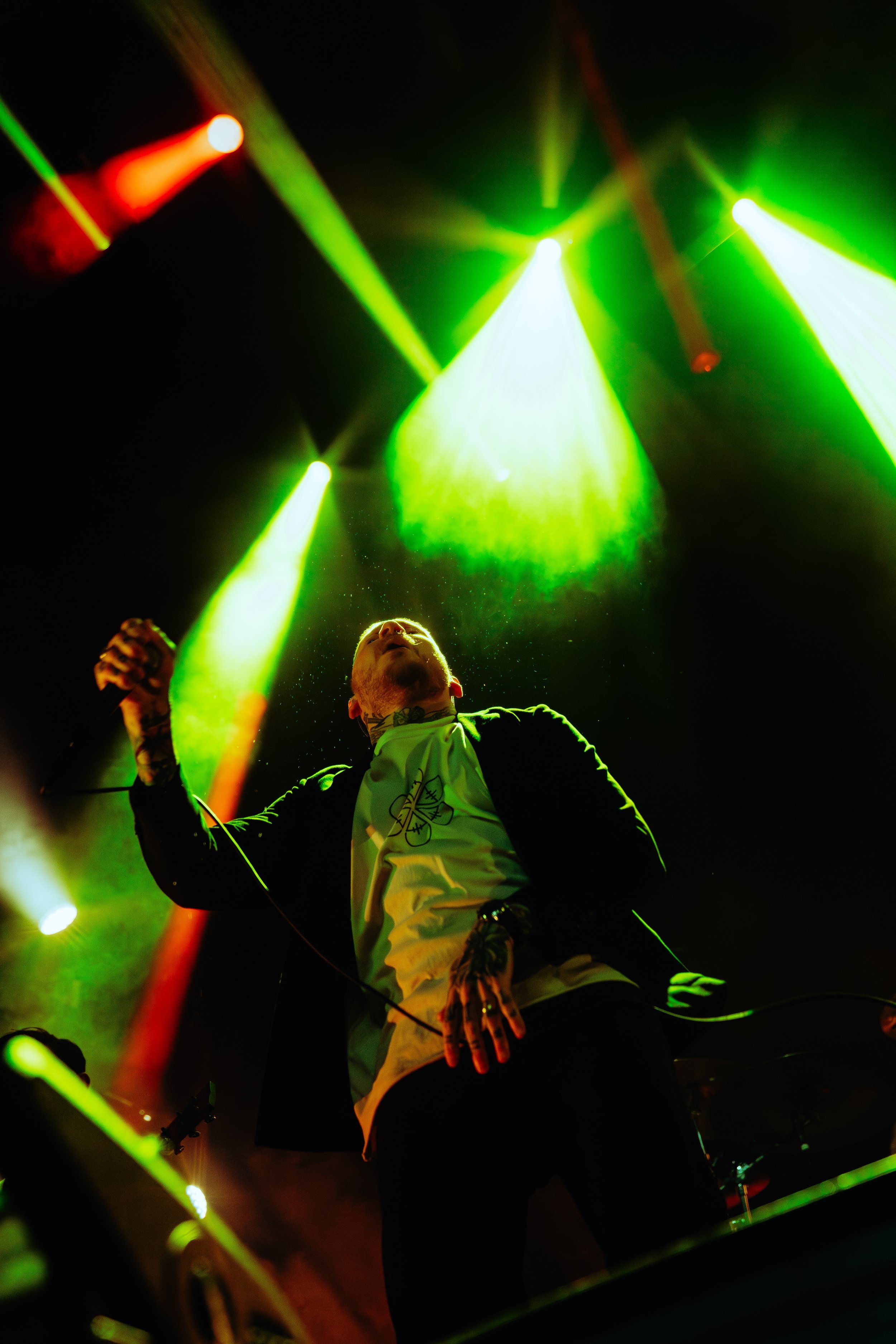 A male singer performing on stage with green and red stage lights shining down on him, holding a microphone in his right hand, and wearing a white shirt with a geometric design.