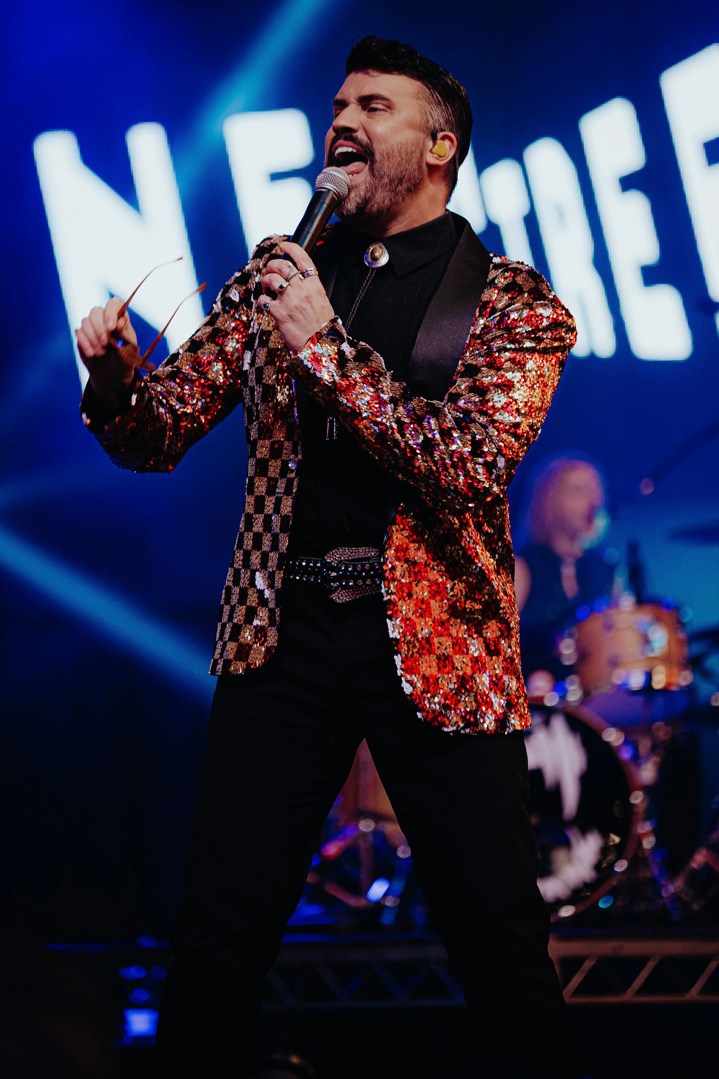 Male performer singing into a microphone on stage wearing a colorful, sequined blazer and black pants, with a yellow in-ear monitor, against a background with blurred lights and a digital display.