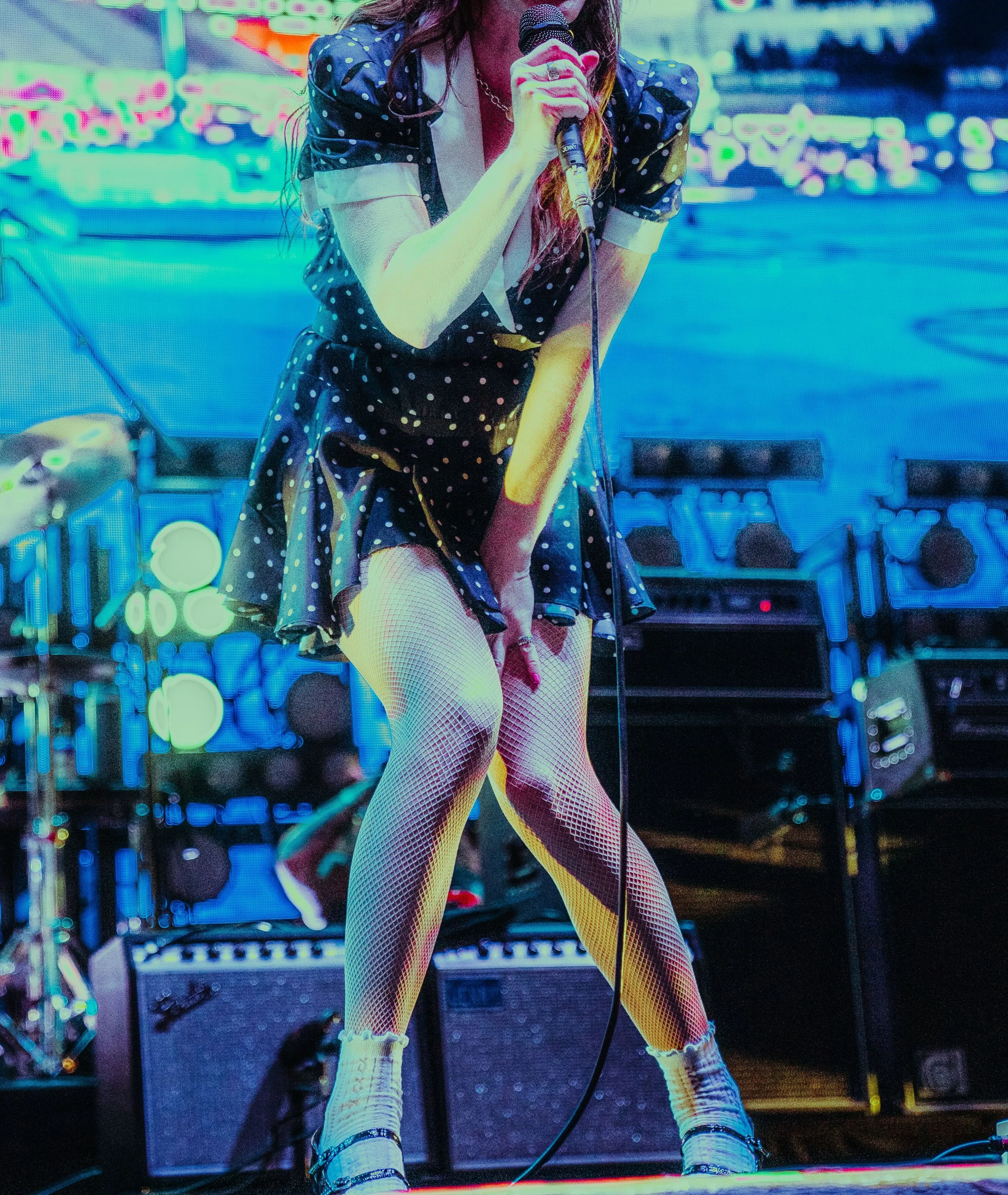 A woman in a polka-dot dress, yellow fishnet stockings, and white socks with black shoes is holding a microphone, performing on stage with colorful, neon-lit background electronics.