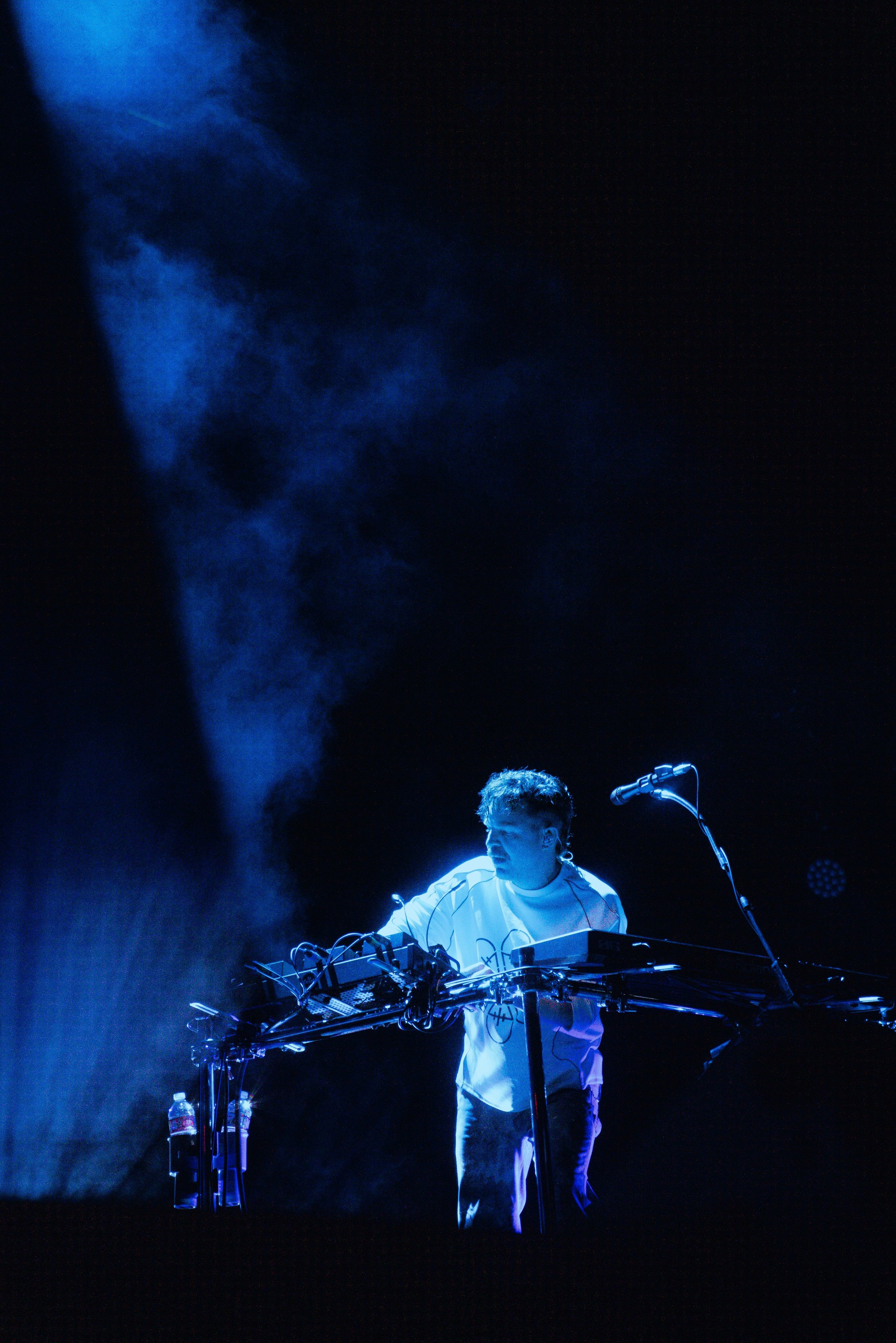 A musician wearing a white shirt performing on stage with blue lighting and fog. He is surrounded by electronic music equipment and a microphone stand.
