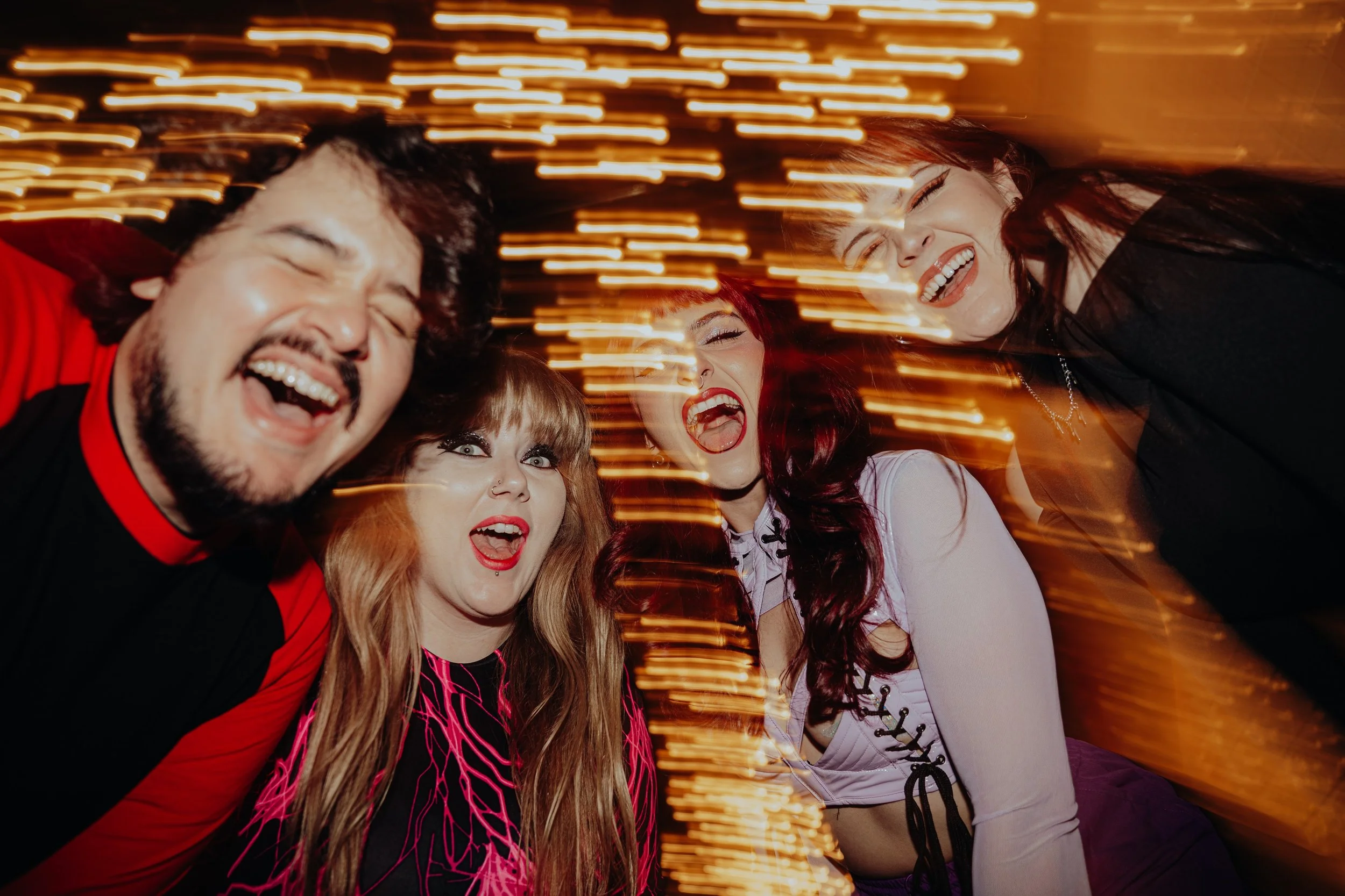 Four friends, two men and two women, laughing and enjoying themselves at a lively party with streaks of warm lights in the background.